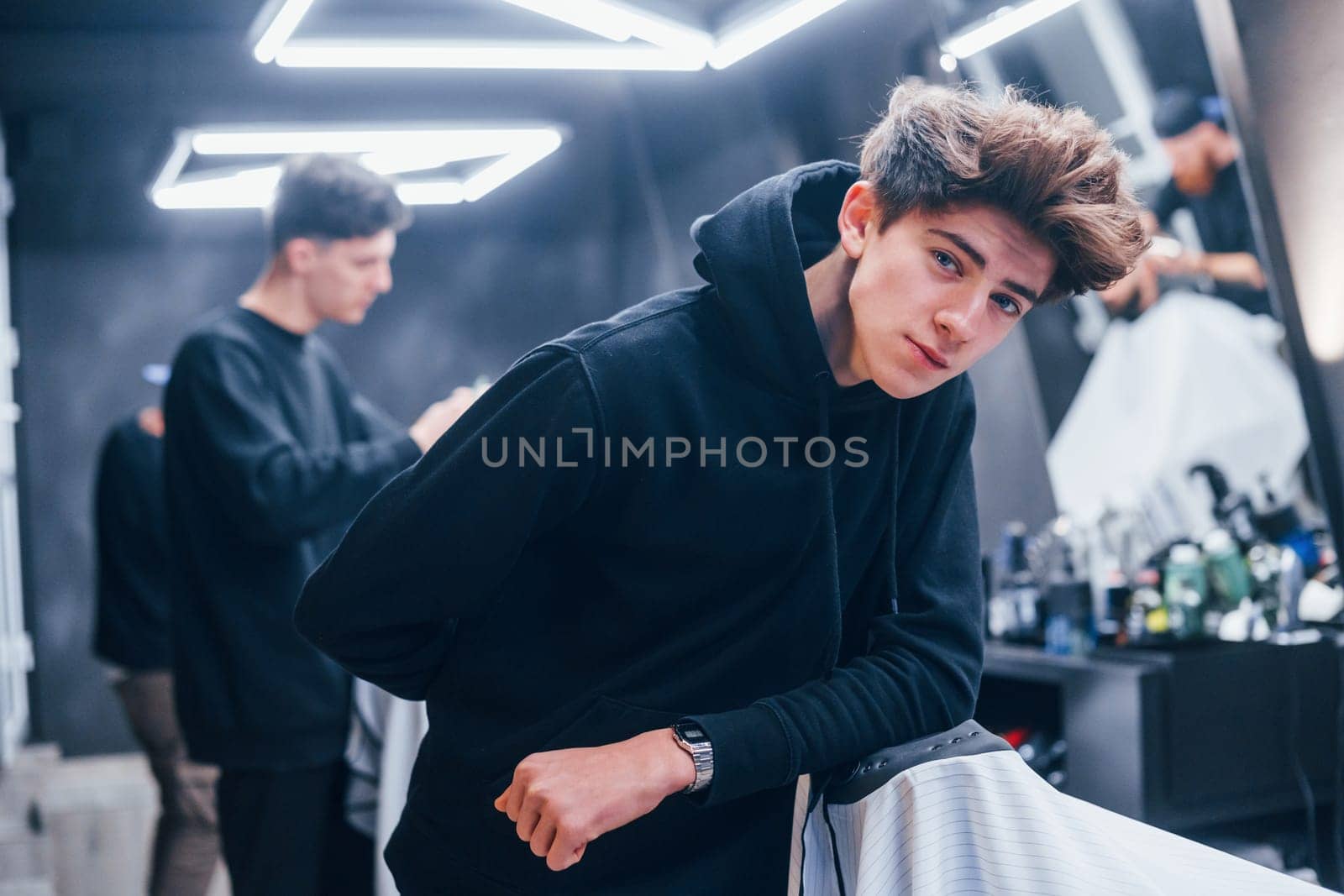 Young barber in black clothes standing in the barbershop. Workers and customer behind.