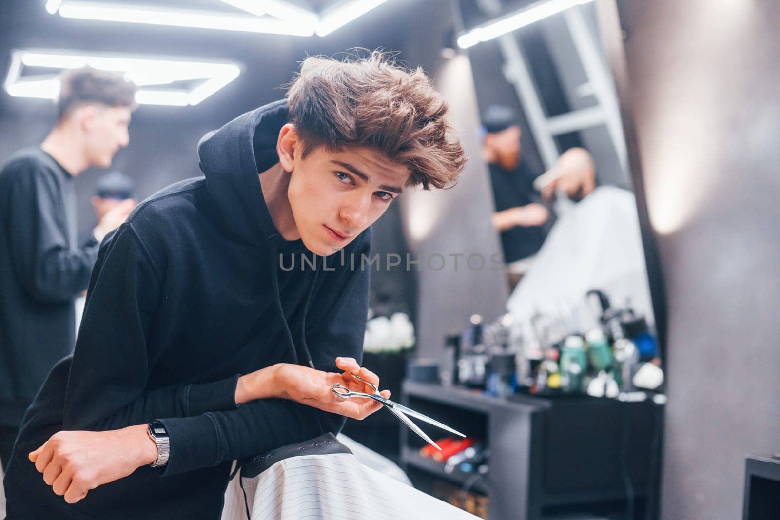 Young barber in black clothes standing in the barbershop. Workers and customer behind.