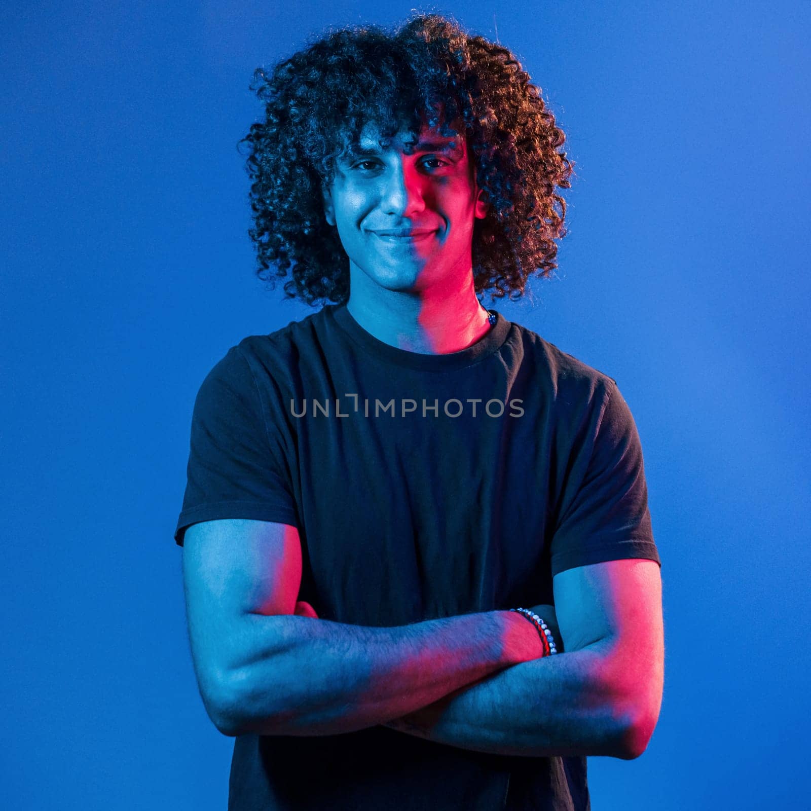 Posing for a camera. Young beautiful man with curly hair is indoors in the studio with neon lighting.