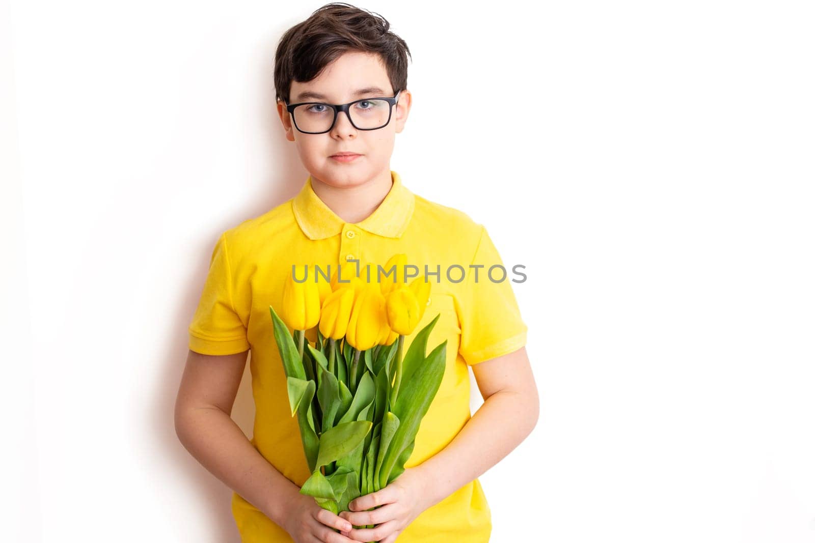 A cute boy in glasses and a yellow T-shirt holds a bouquet of yellow tulips, stands on a white background, isolated. Close up. Copy space.