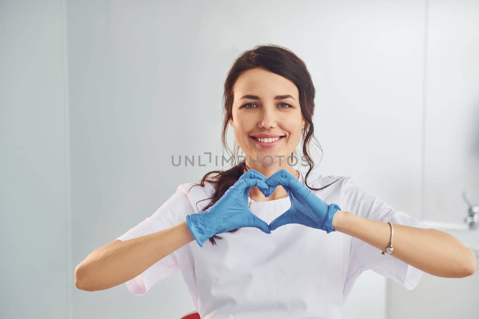 Heart shaped gesture. Portrait of professional female dentist with equipment that standing indoors.