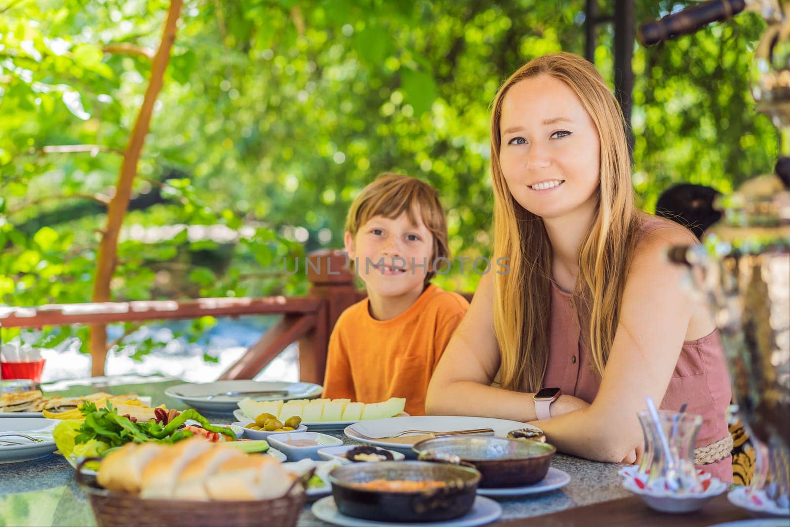 Mom and son eat Turkish breakfast. Turkish breakfast table. Pastries. Vegetables. Olives. Cheeses, fried eggs. Jams, tea in copper pot and tulip glasses. Wide composition.