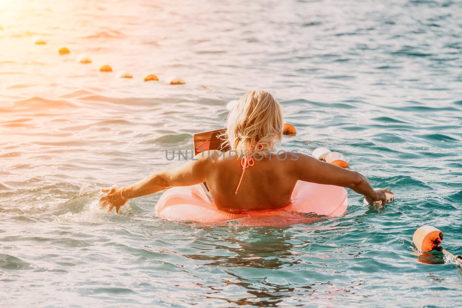 Woman works on laptop in sea. Freelancer, young blond woman in sunglases floating on an inflatable big pink donut with a laptop in the sea at sunset. Freelance, travel and holidays concept. by panophotograph