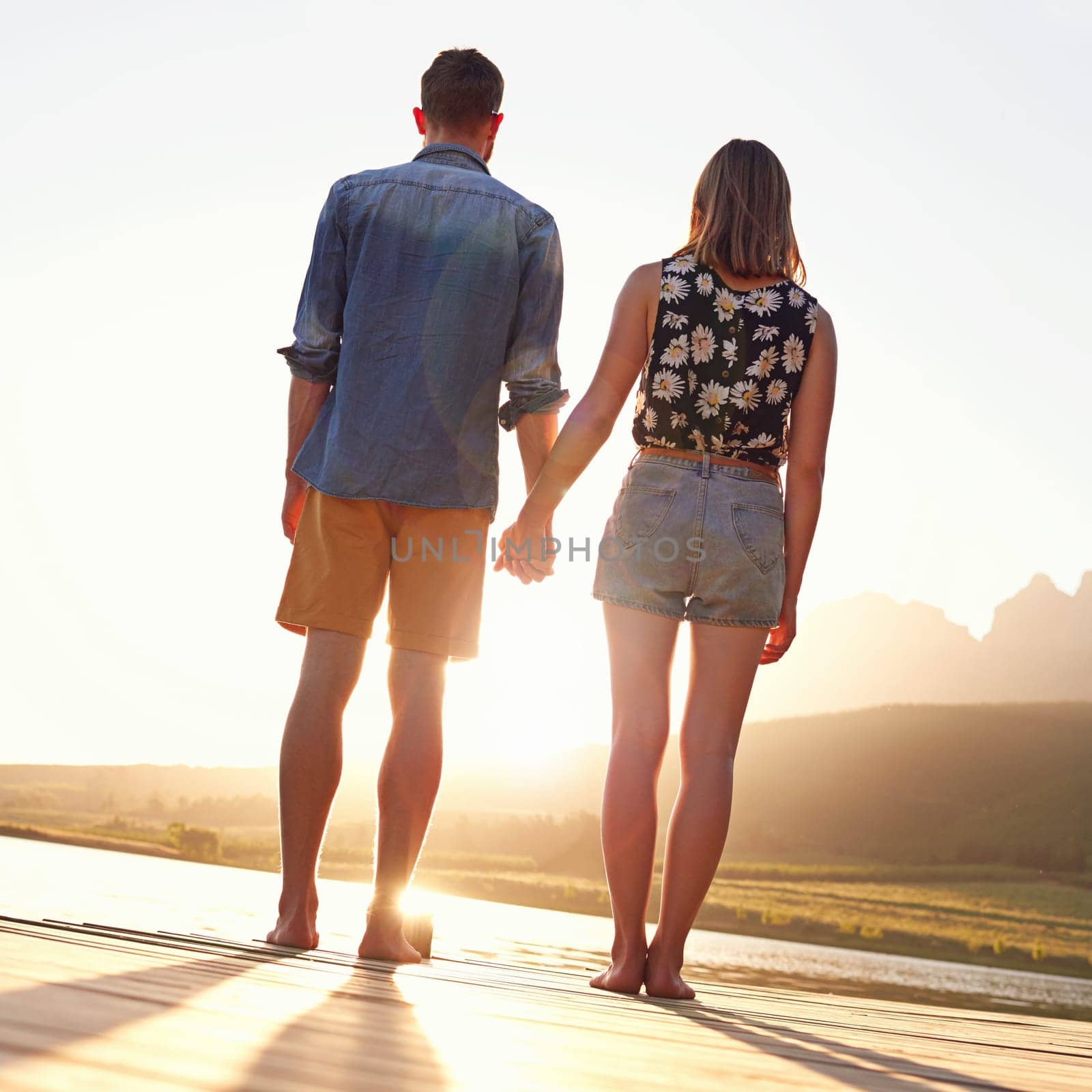 Love and sunsets. Rearview shot of an affectionate young couple standing on a dock at sunset. by YuriArcurs