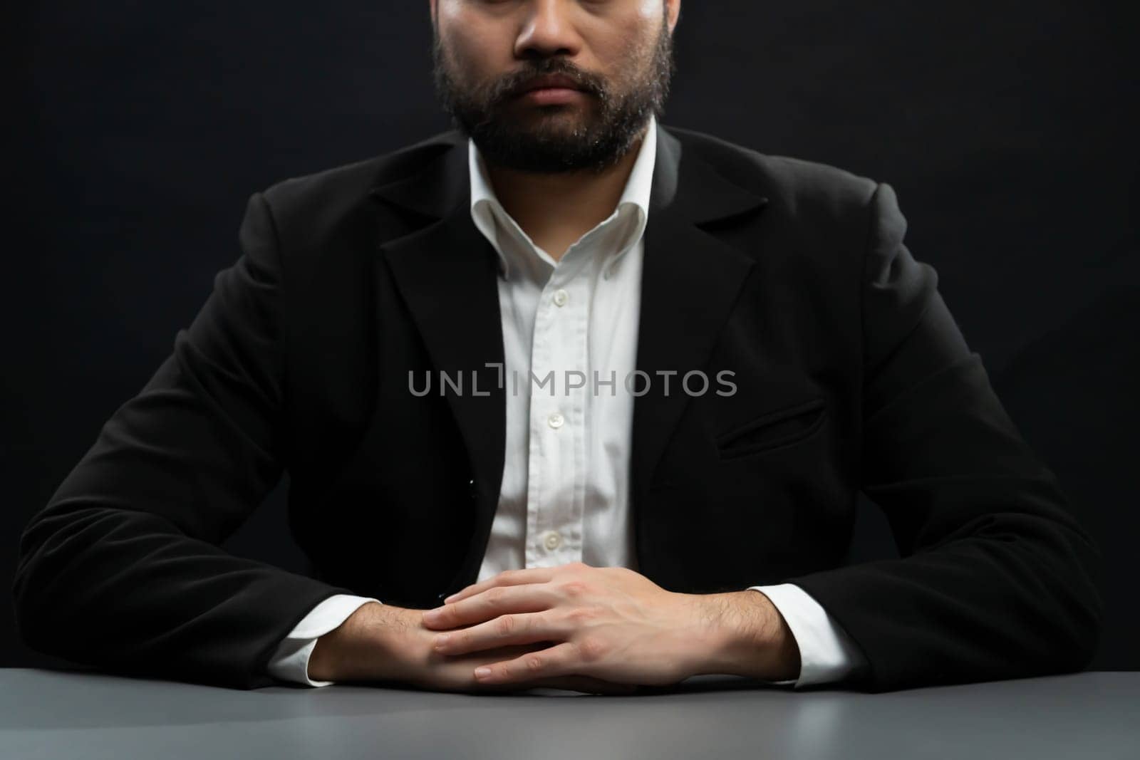 Businessman or lawyer wearing formal black suit sitting at table on isolated black background. Concept of a man with authority and seriousness gesture. equility