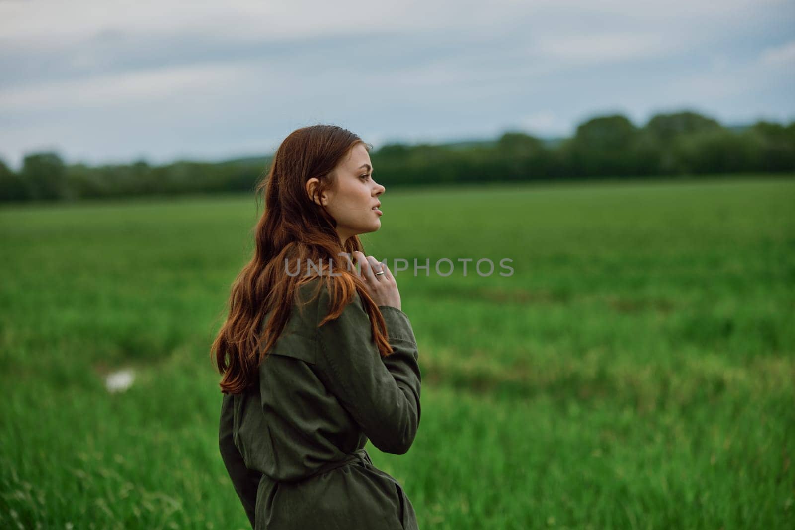 a beautiful, red-haired girl in a raincoat stands in a field in the spring in rainy weather. High quality photo