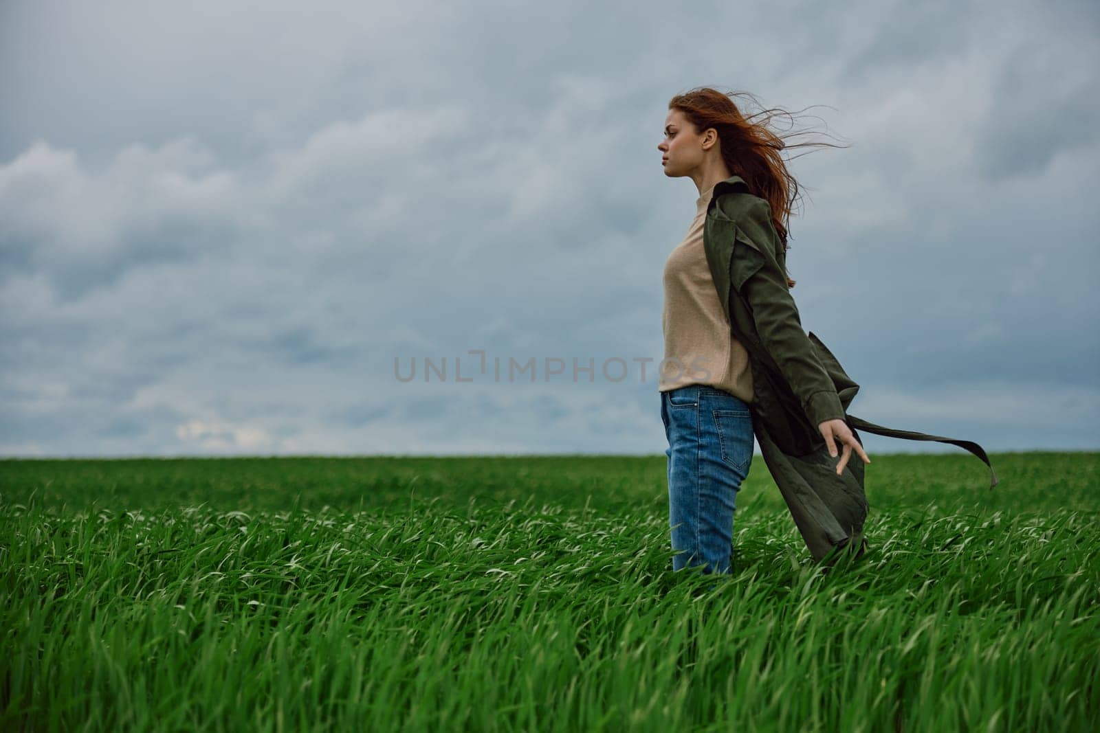 a red-haired woman in a long coat stands in a green field and the wind blows her hair. High quality photo