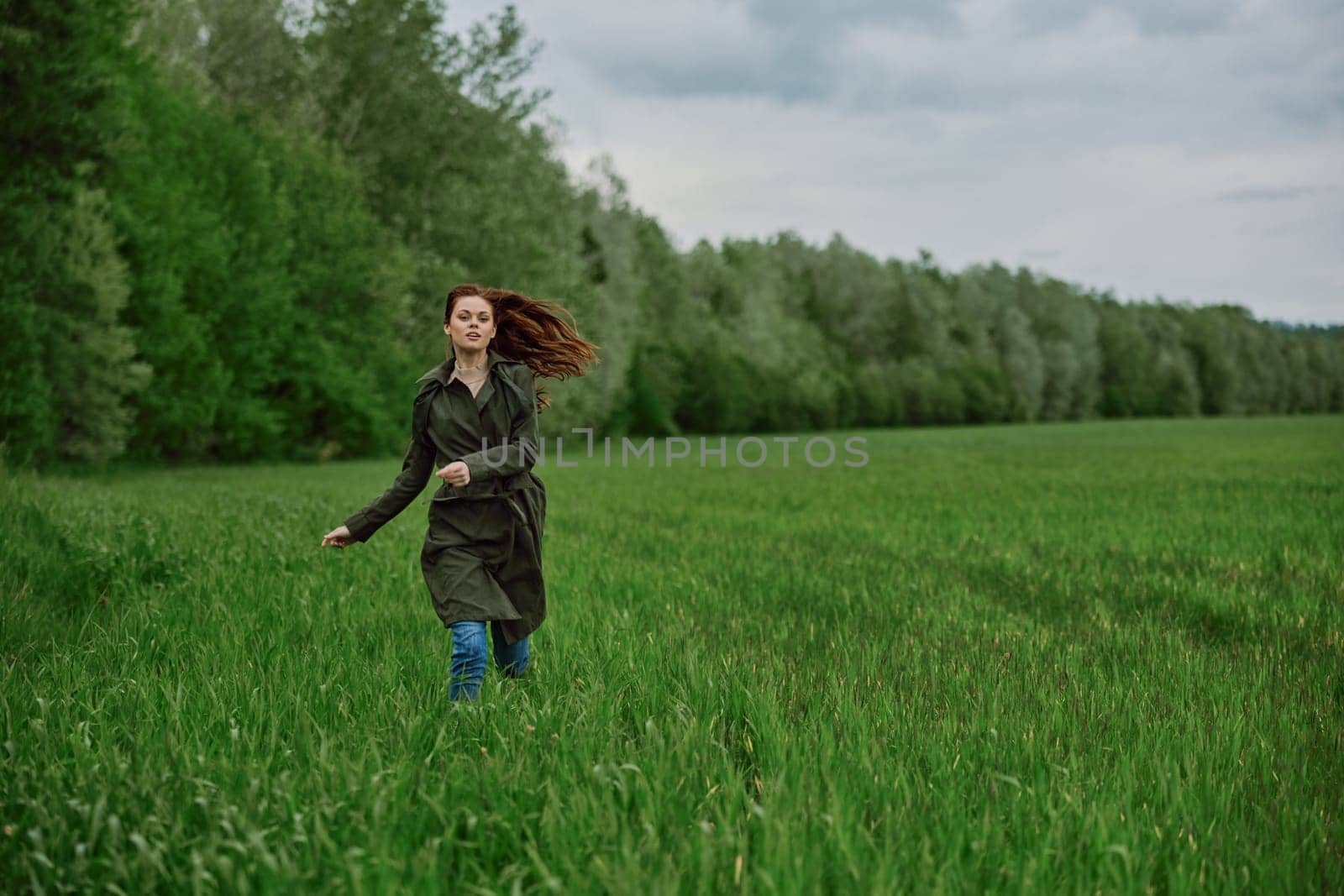 a beautiful woman in a long raincoat runs across a field in high grass in spring in cloudy weather. High quality photo