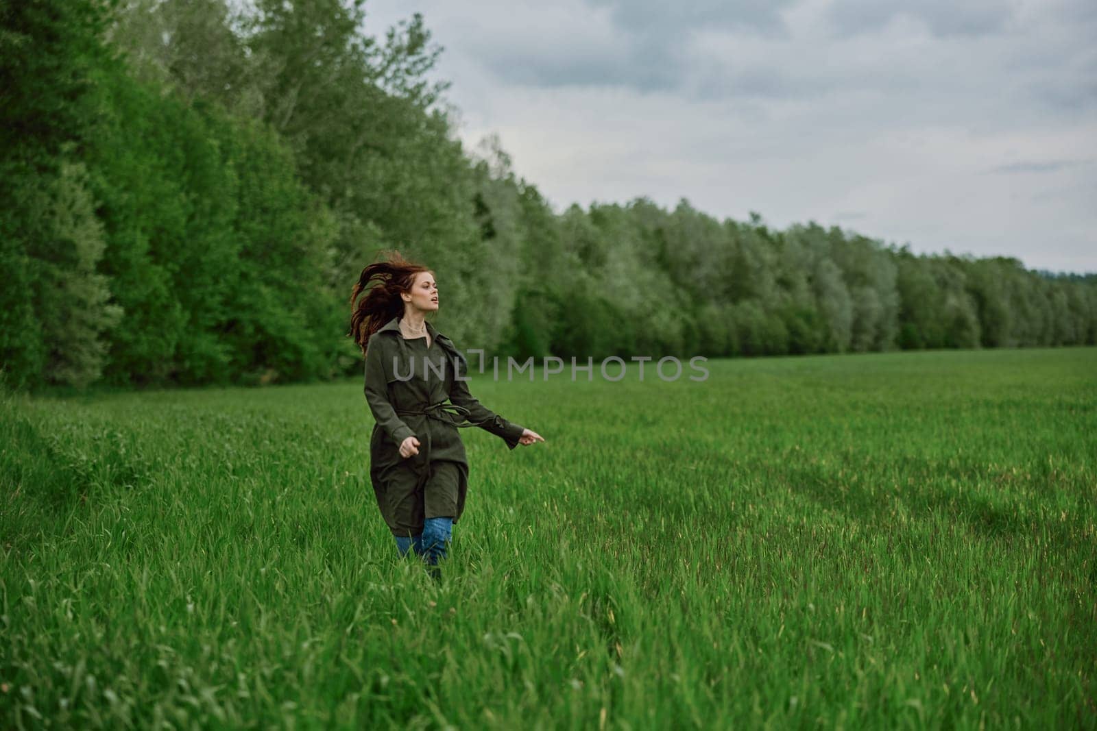 a beautiful woman in a long raincoat runs across a field in high grass in spring in cloudy weather. High quality photo