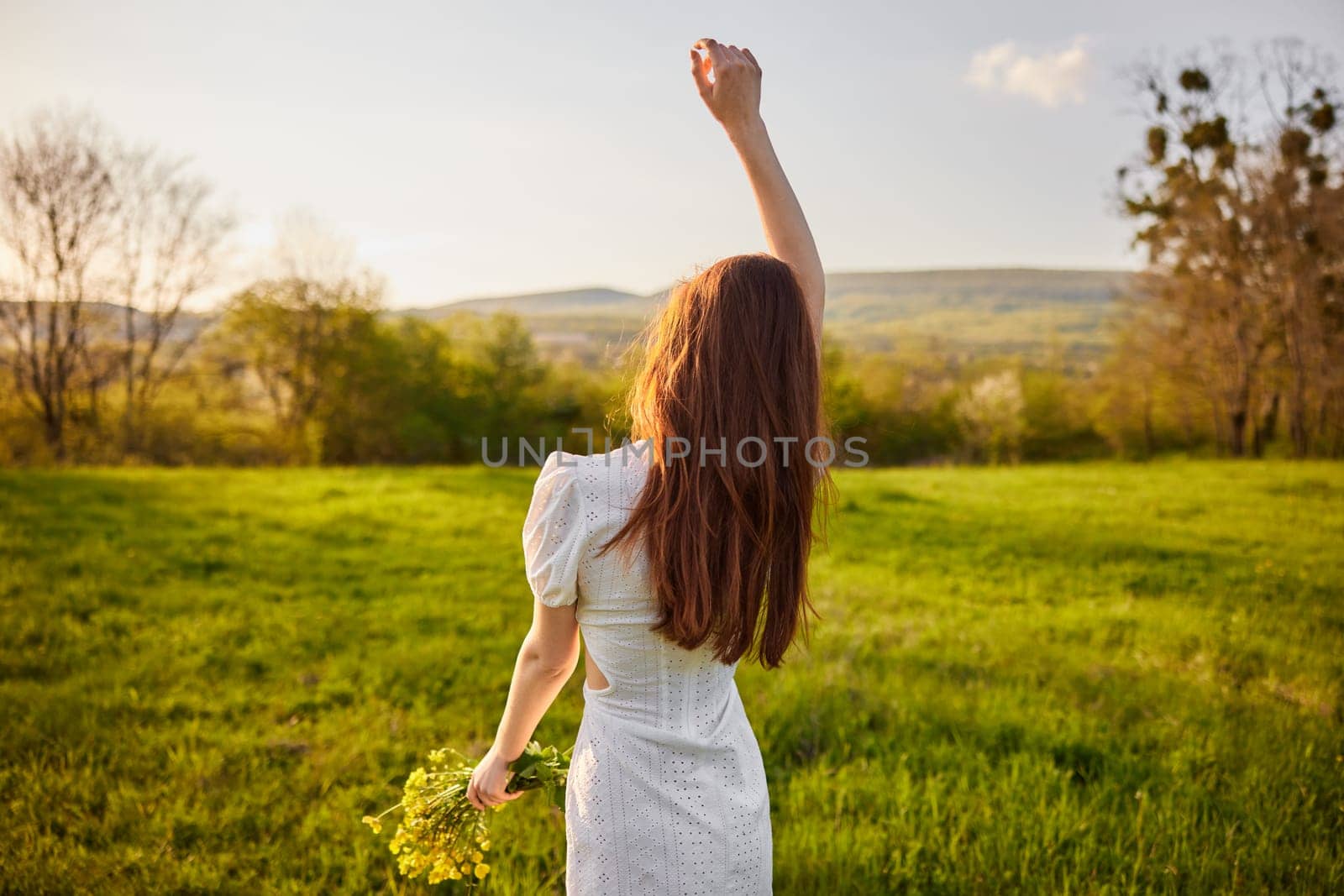 photo from the back of a red-haired woman raising her hands in happiness with a bouquet of flowers in the rays of the setting sun. High quality photo