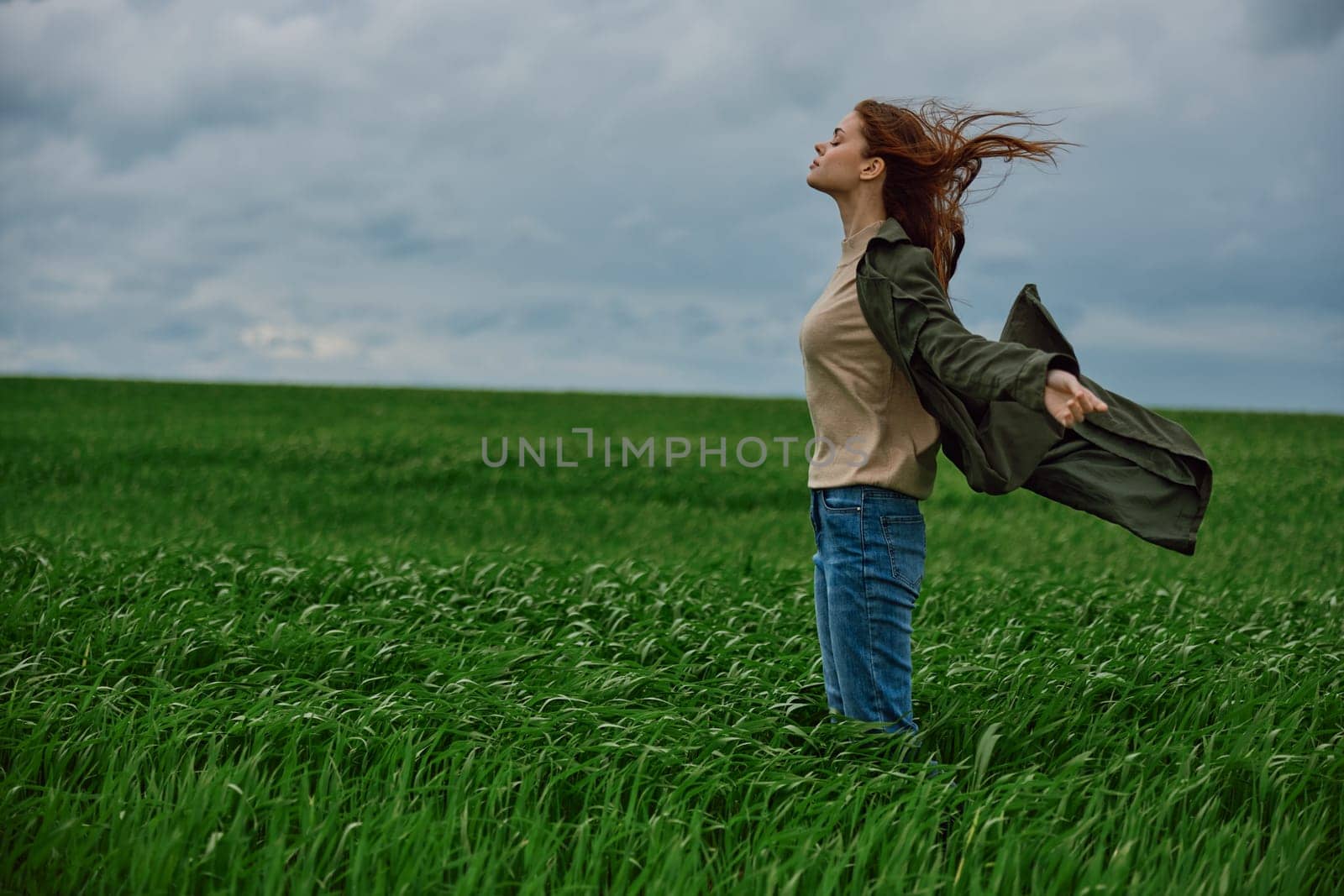a red-haired woman in a long coat stands in a green field and the wind blows her hair by Vichizh