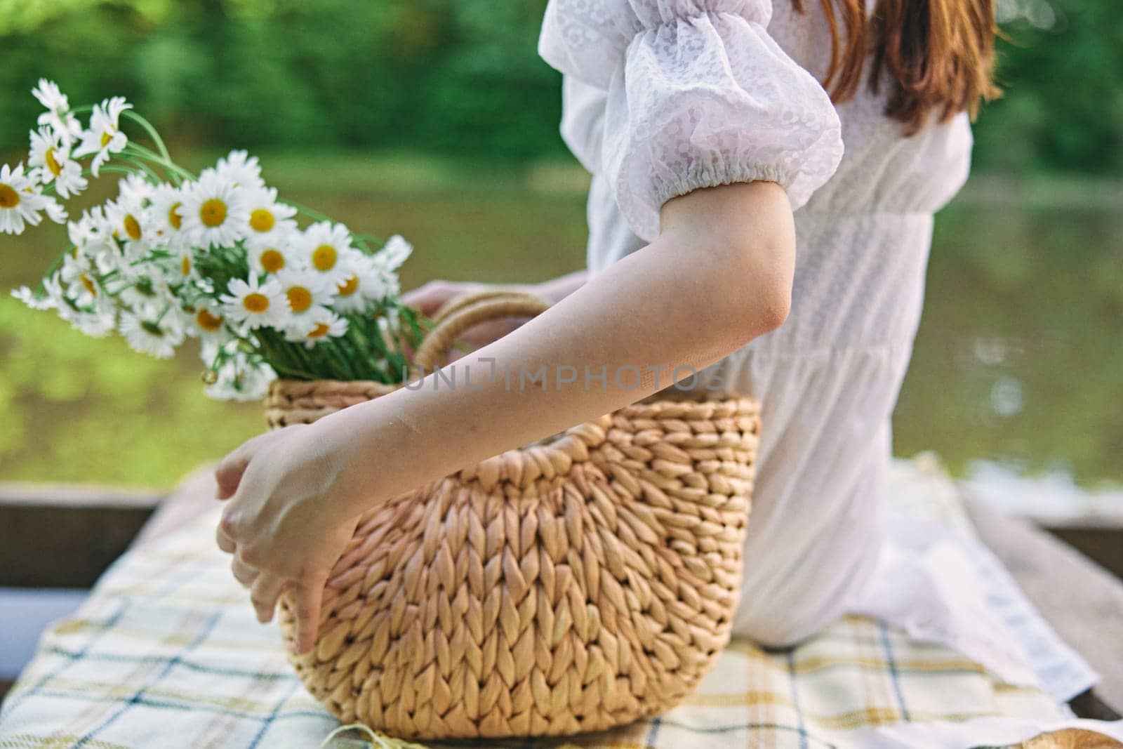 a woman sits with her back to the camera by the lake holding a wicker basket with daisies in her hand. subject photography without a face by Vichizh