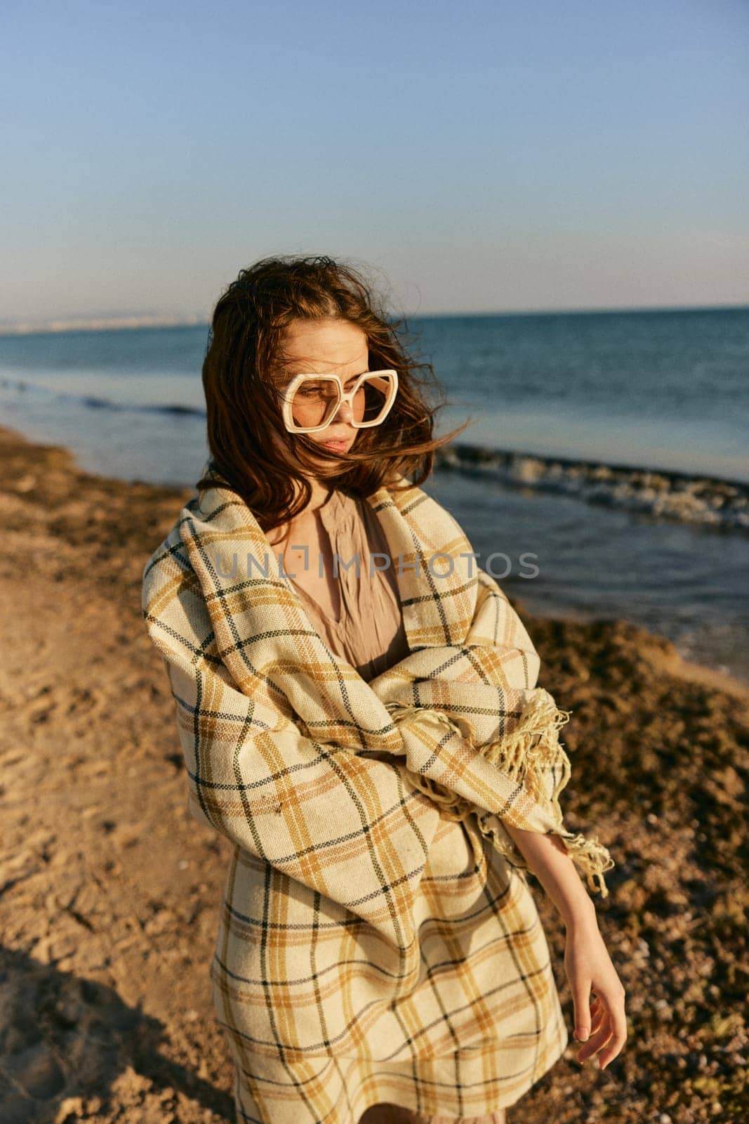 a woman in large glasses wrapped in a plaid enjoys the view of the setting sun standing on the beach. High quality photo
