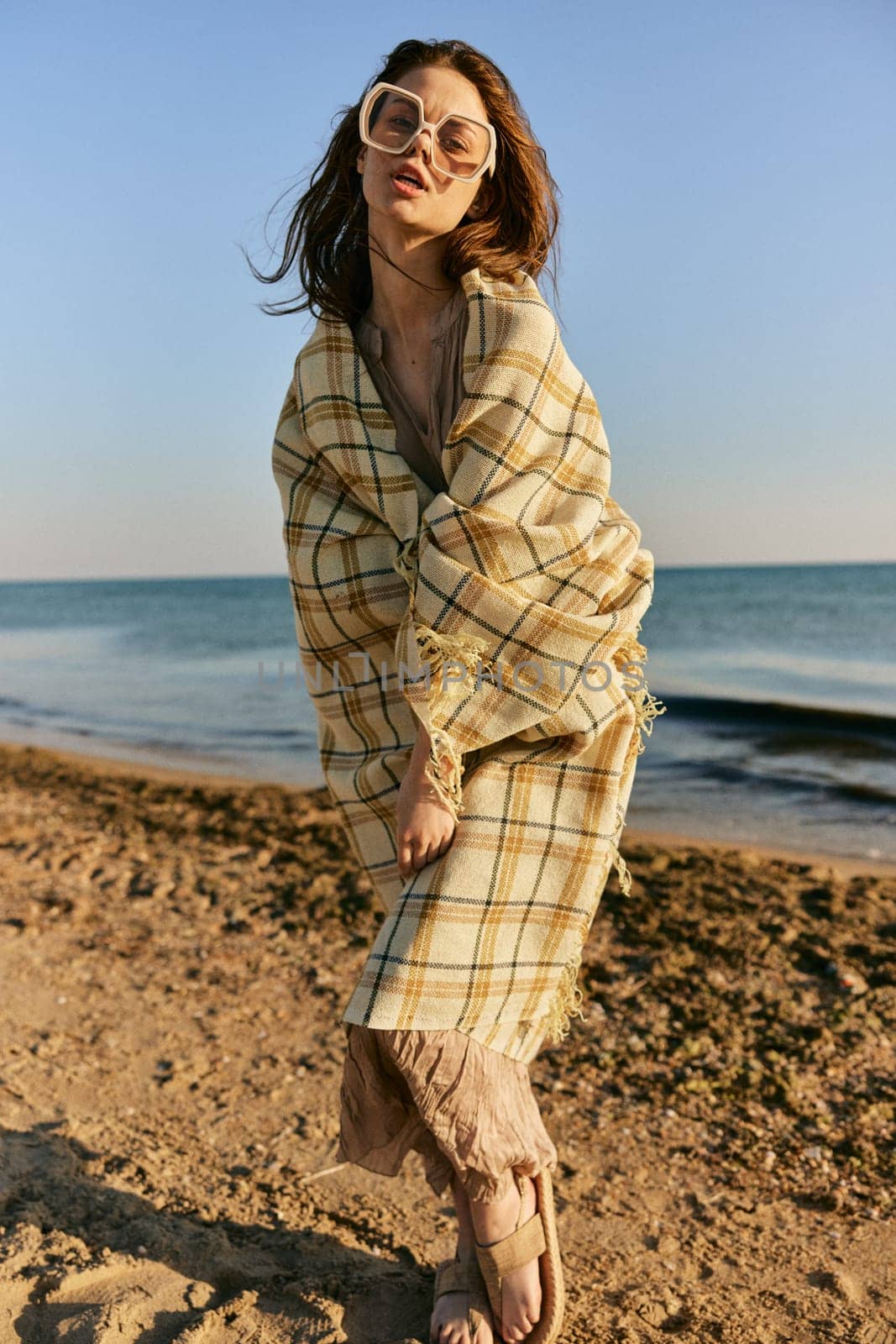 a woman wrapped in a plaid stands on the seashore in bright sunglasses and squints from the sun. High quality photo