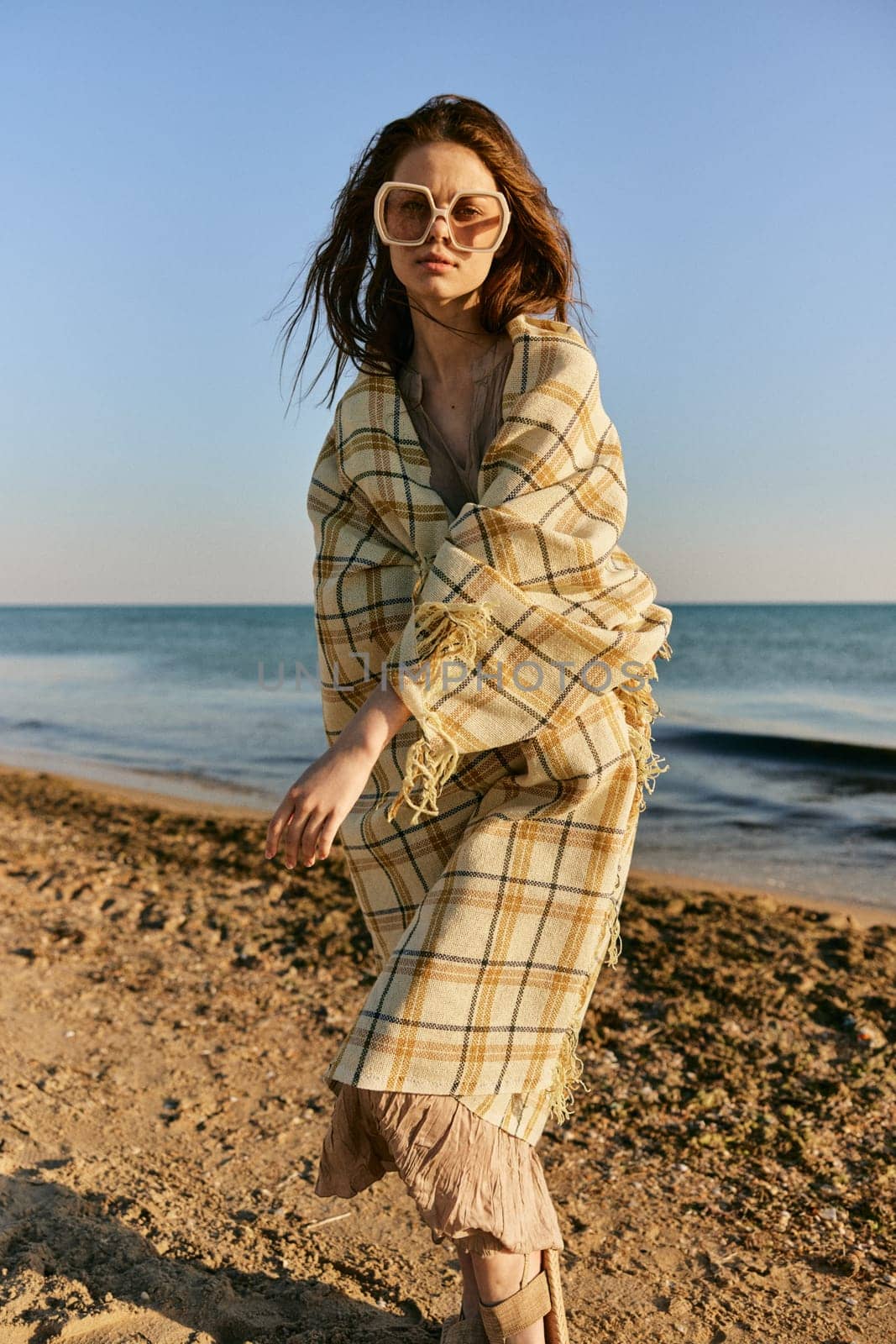 a woman wrapped in a plaid stands on the seashore in bright sunglasses and squints from the sun. High quality photo