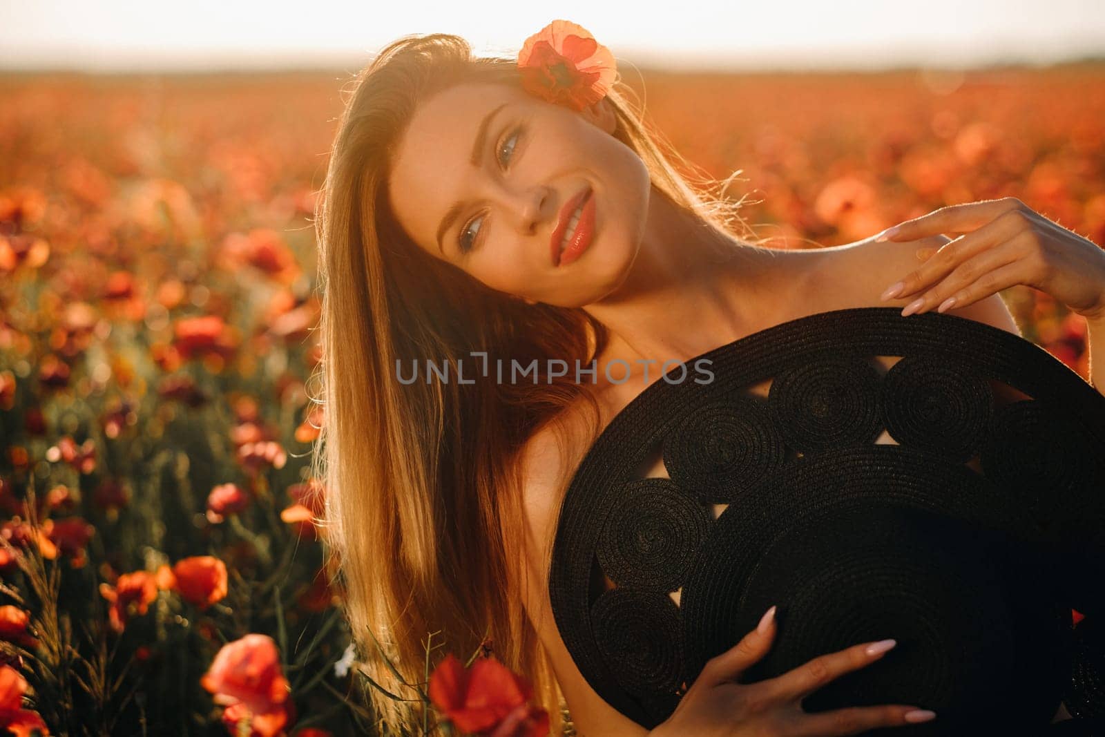 naked girl with a black hat in her hands in a poppy field at sunset.