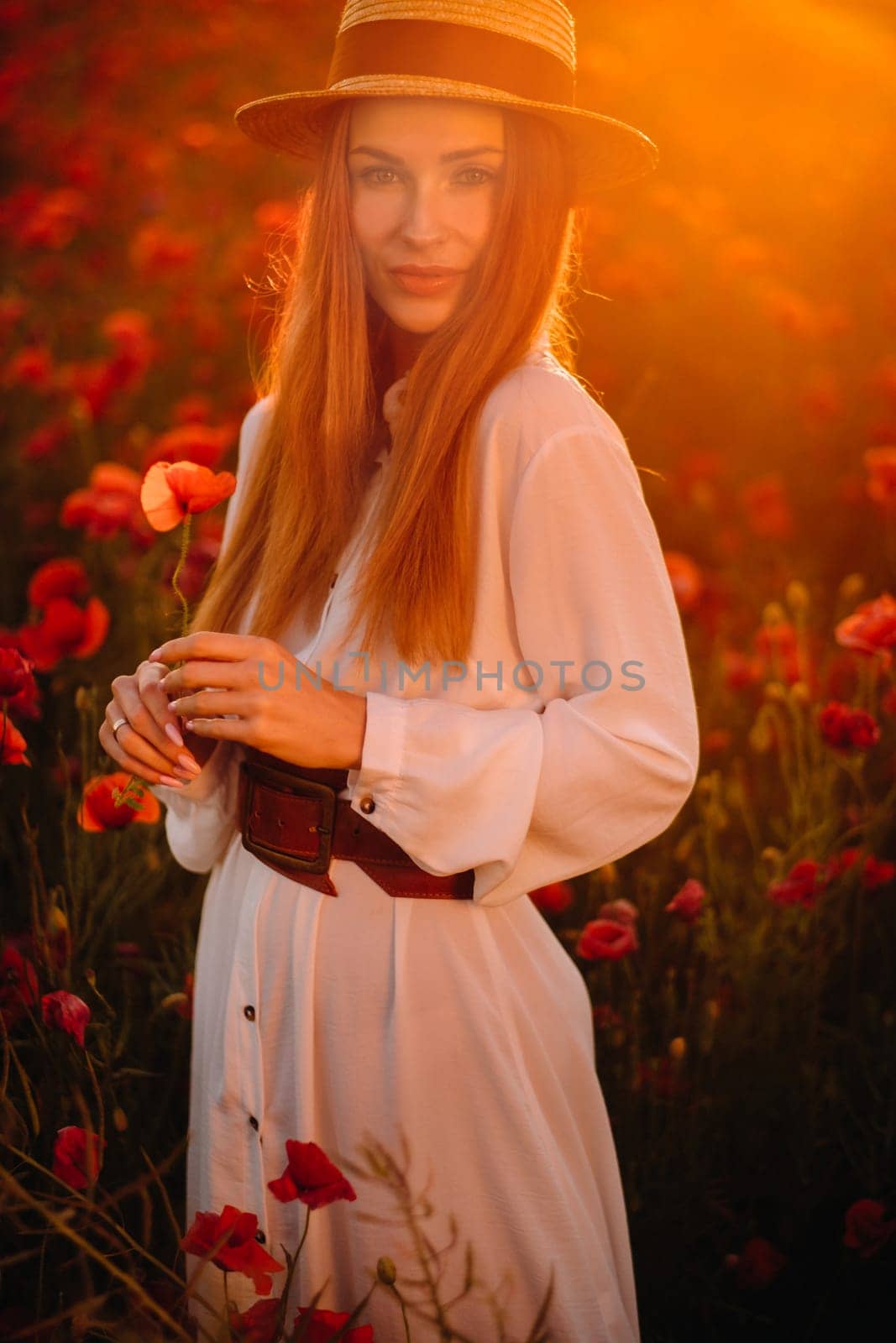 a girl in a white dress and hat stands in a field with poppies at sunset and holds a poppy flower in her hand.
