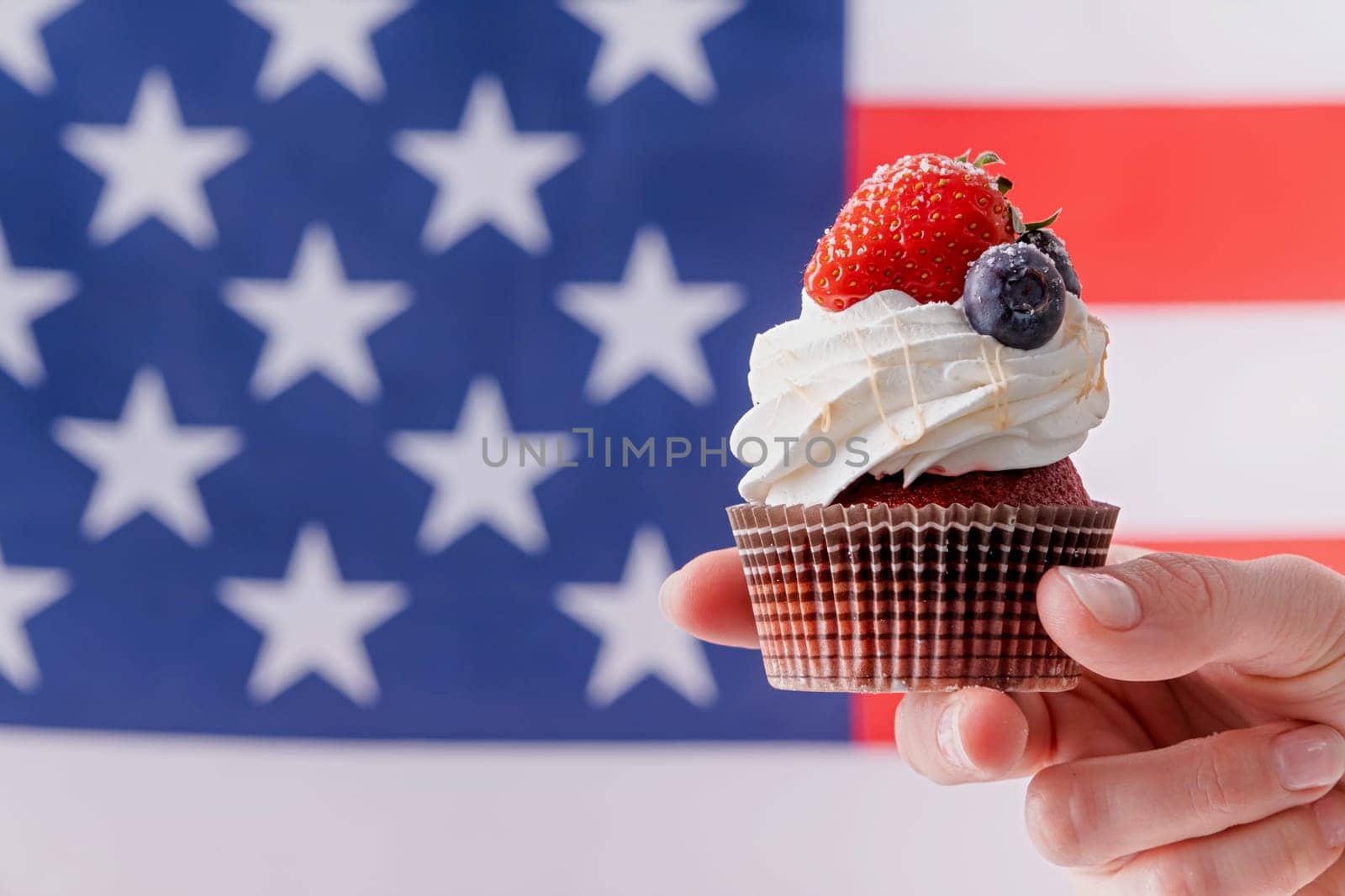 hand holding sweet cupcake with blueberries and strawberry , flag background by Desperada