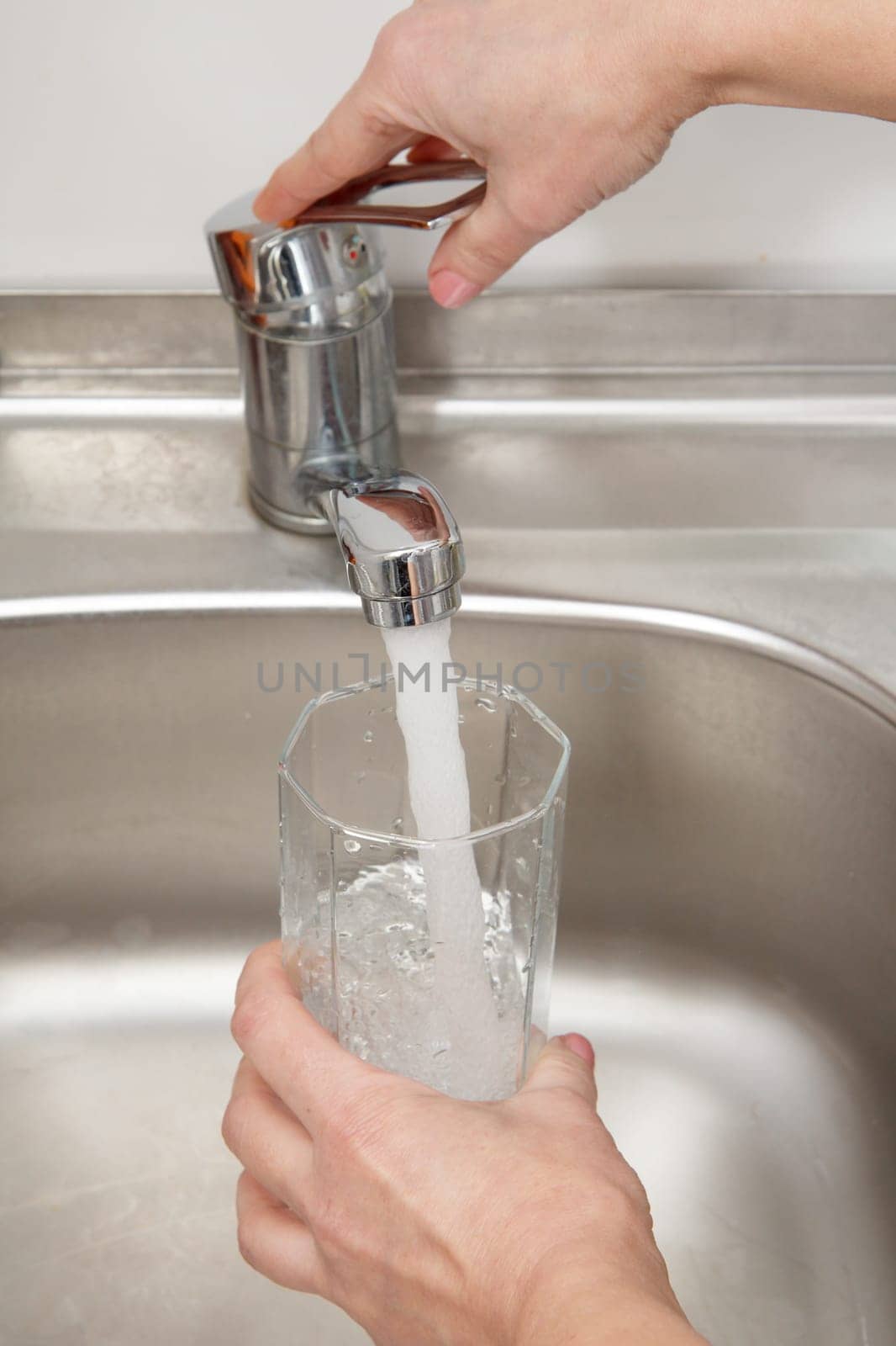 Glass in female hand is filled with clean transparent water under the tap with metal sink on the background