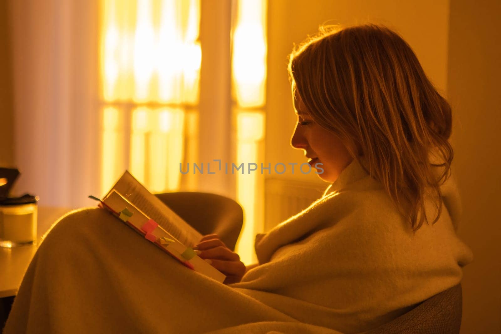 Smiling young beautiful woman reading a book, wrapped in a blanket while sitting in the living room at sunset light.