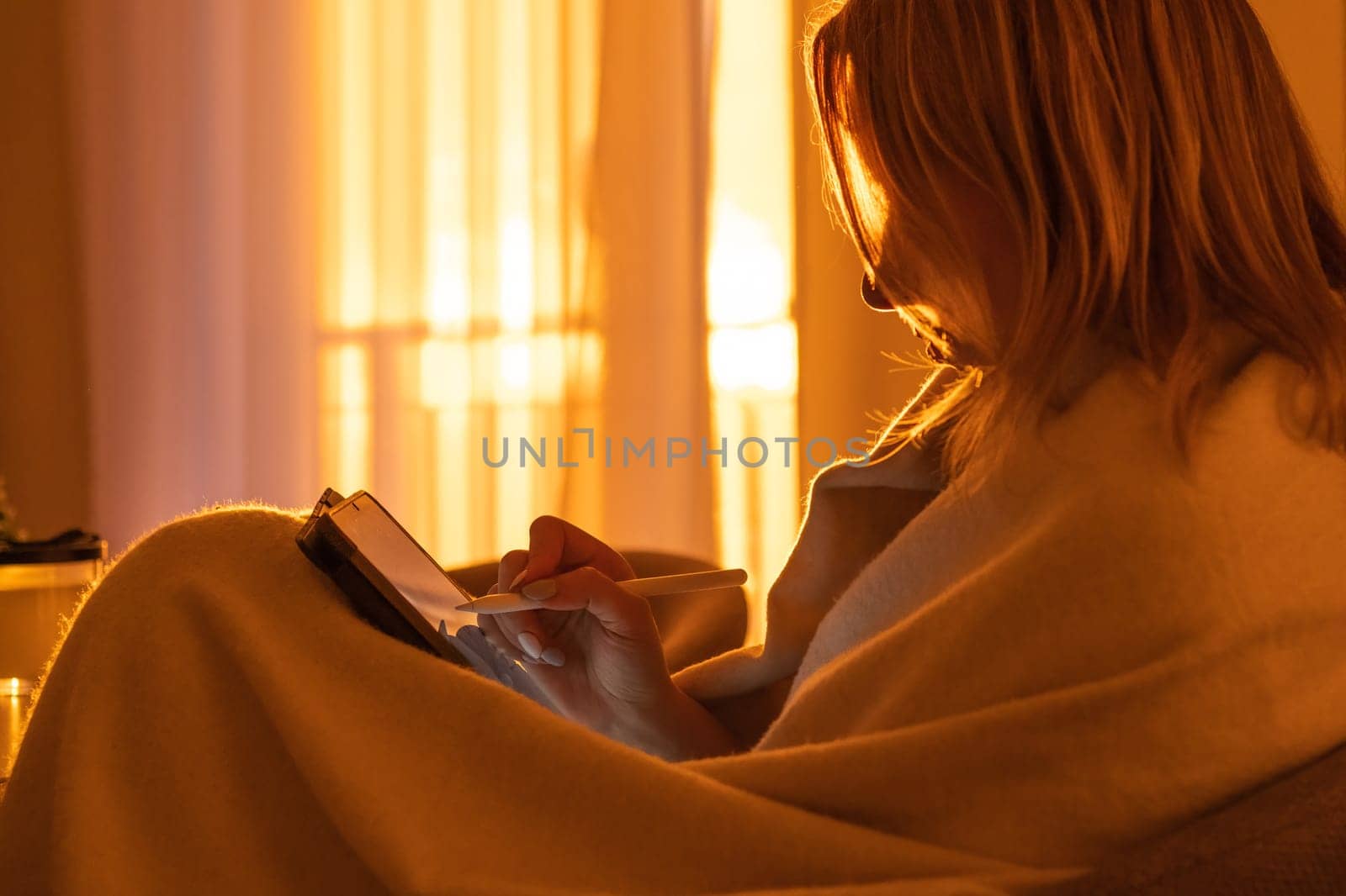 Woman wrapped in the blanket and uses a tablet while sitting in the living room at sunset light by vladimka