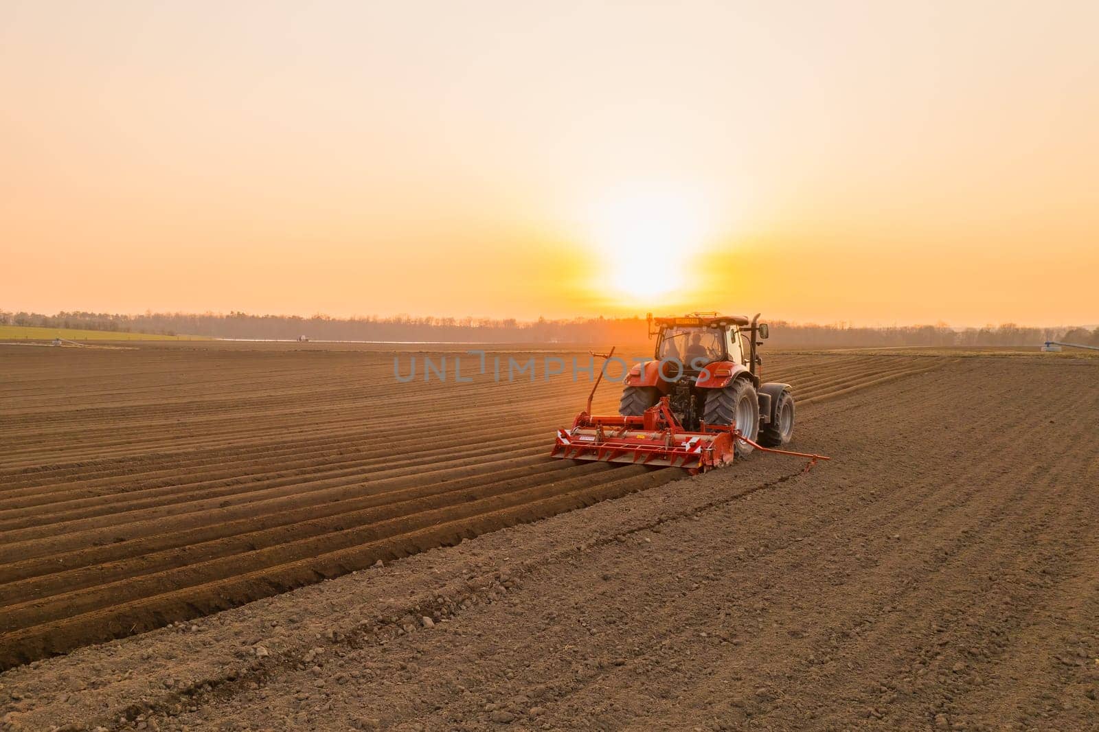 PRAGUE , CZECH REPUBLIC - MARCH 18 2022: Modern tractor cultivates soil in field on agricultural farm at bright sunset. Powerful machine works dragging plow behind at rural site in evening