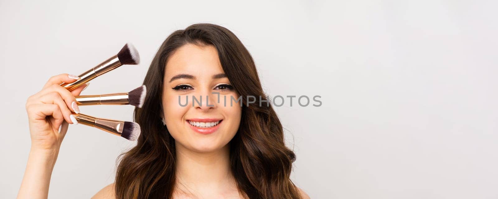 Closeup portrait of a young smiling woman with makeup brushes near her face on the grey background with copy space.