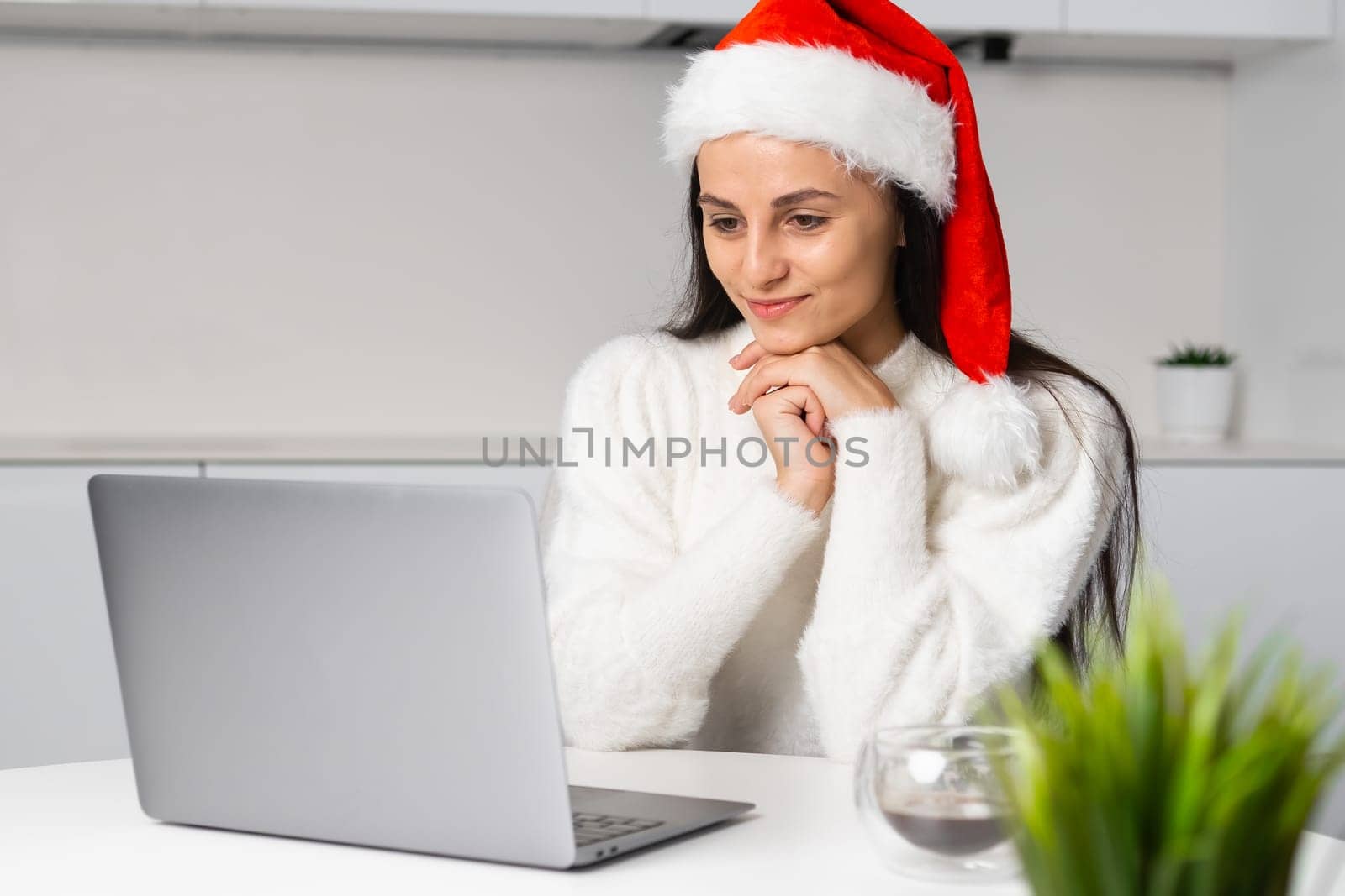 A charming woman in a Santa Claus hat has an online meeting on Christmas holidays at modern home interior.