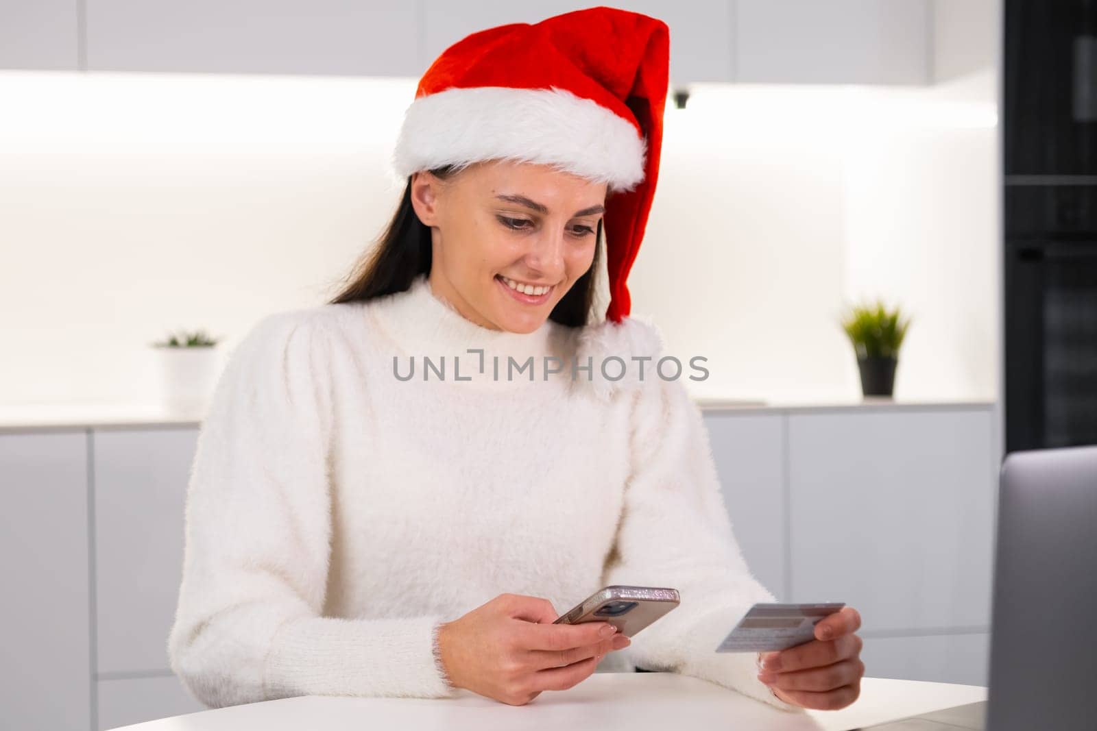 A happy woman in a red Santa Claus hat uses a credit card and smartphone for online shopping at home.