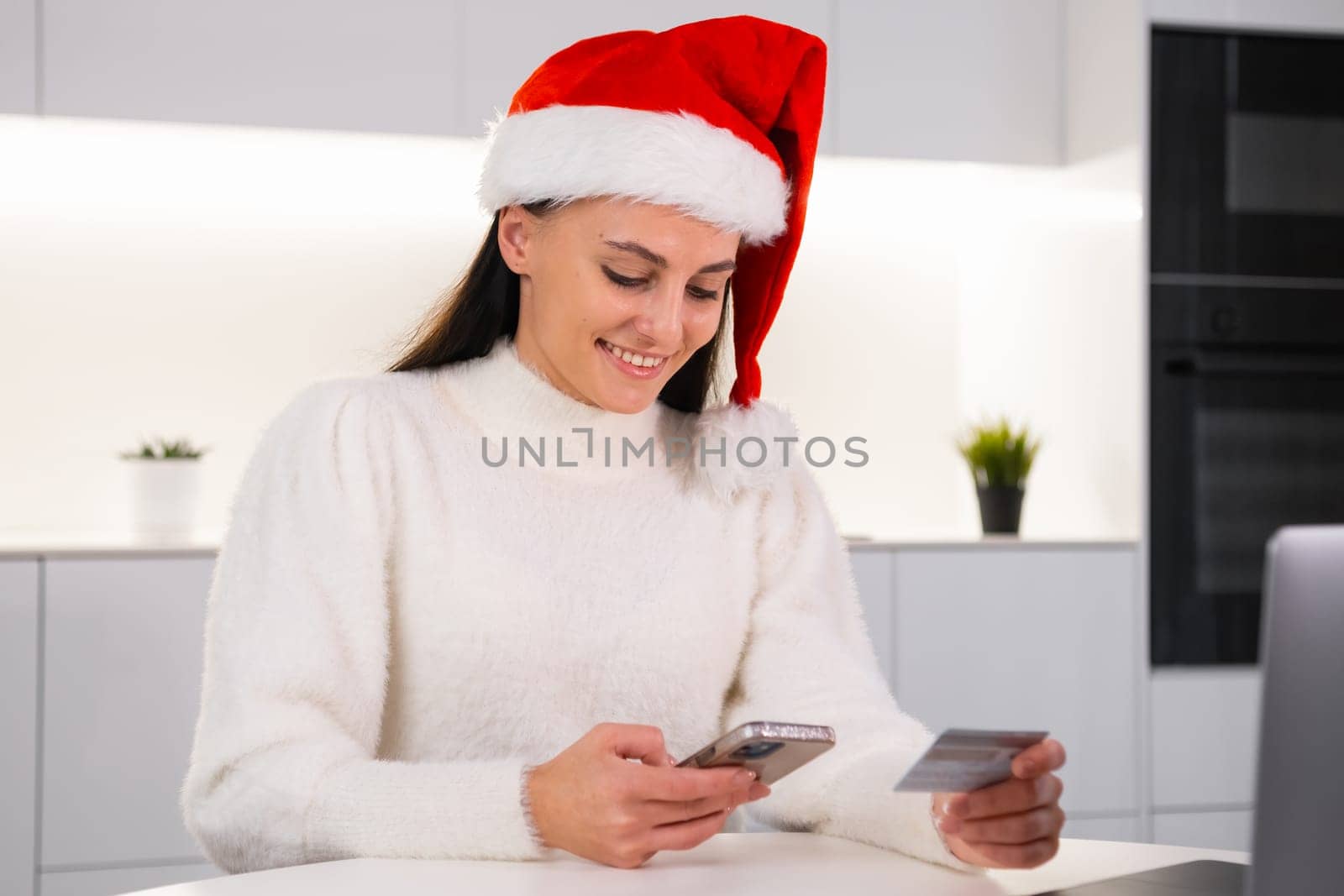 Happy woman buying gifts online sitting in the modern kitchen. Woman in Santa Claus hat spending money on sale via the internet using a credit card.