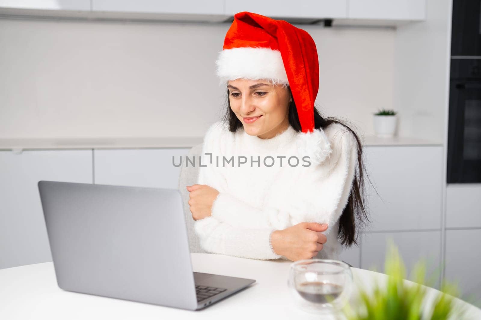 A smiling woman in a red Santa Claus hat has a video chat on a laptop sitting in the kitchen. Online meeting at home.