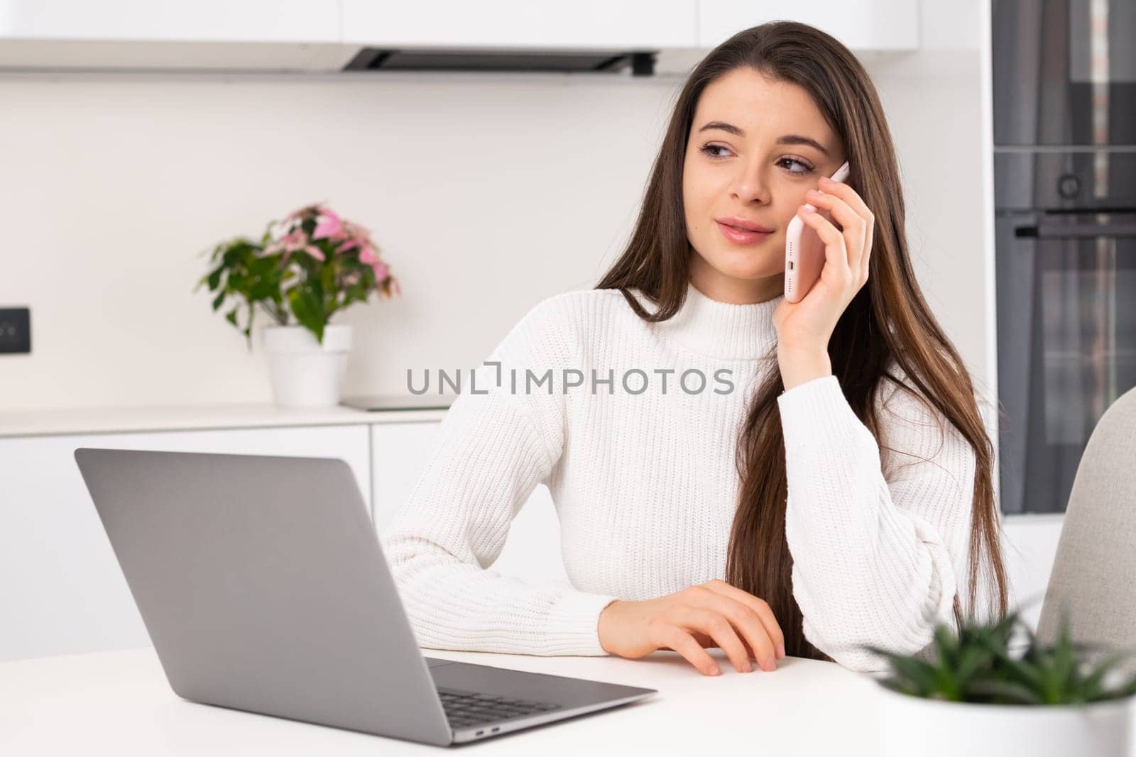 Female freelancer calls to friend on smartphone discussing life at break. Young woman talks on phone sitting at table near Notebook