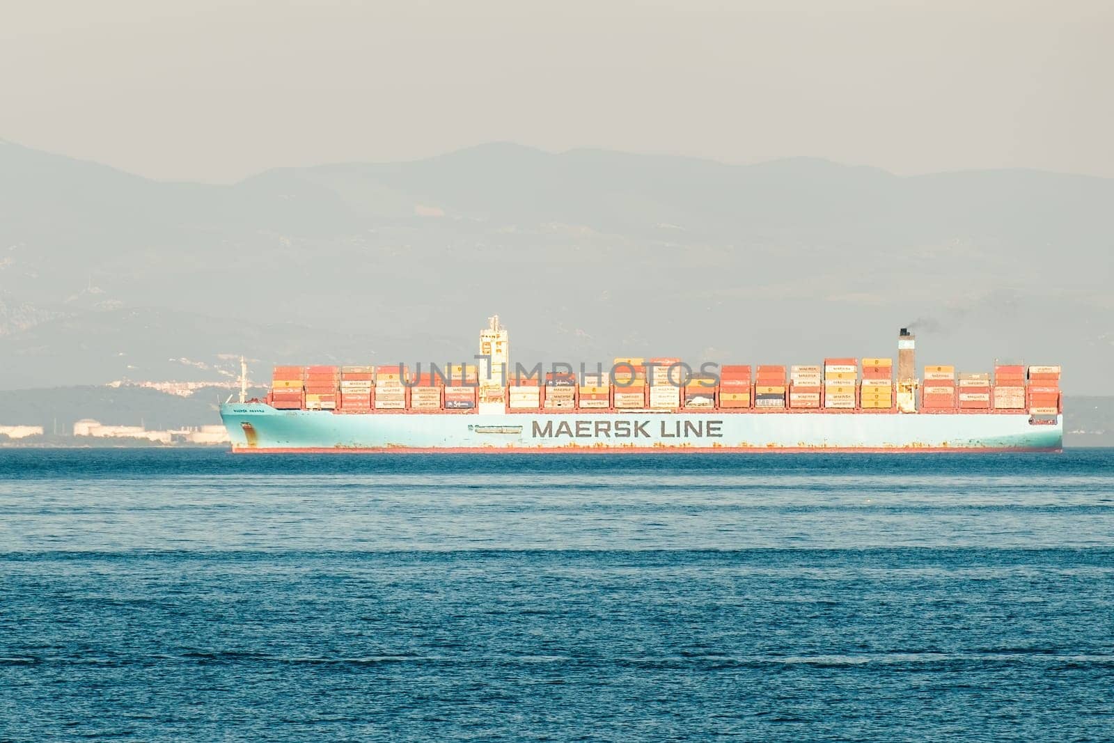 RIJEKA, CROATIA - JULY 10 2022: Container ship sails on deep blue ocean. White vessel transports containers against blurry dull mountain on coast and overcast sky on July 10 in Rijeka