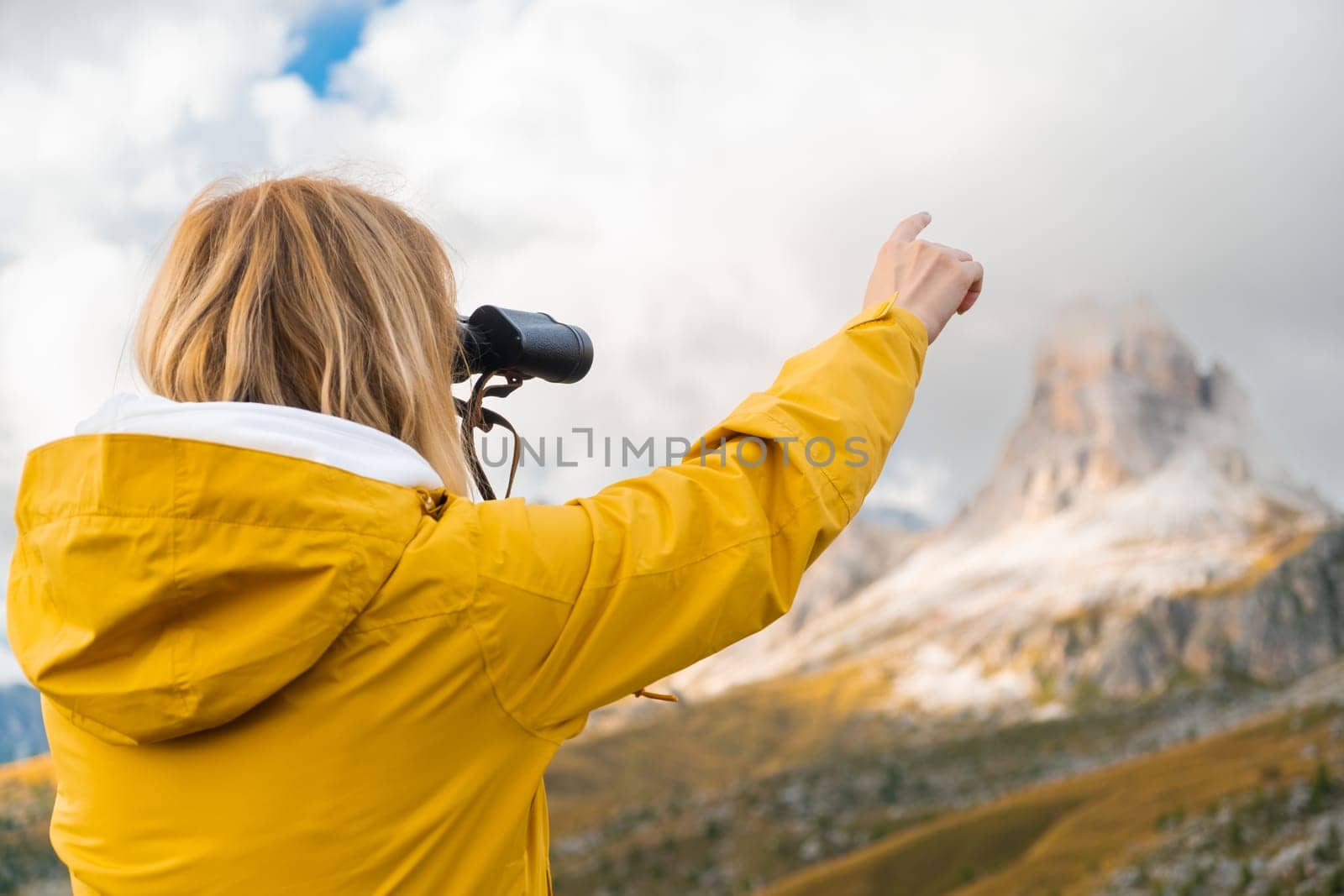 Blonde woman looks at Passo Giau pass through binoculars pointing finger on rocky mountain. Female tourist enjoys hiking in Italian Alps slow motion