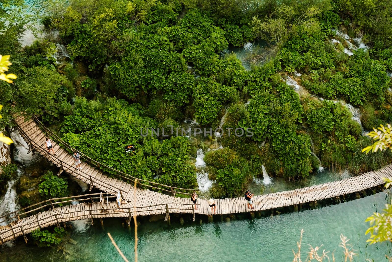 Hiking tourists walk on wooden footbridge surrounding lake in natural reserve. Majestic view of blue lake among picturesque landscape on Plitvice lakes upper view