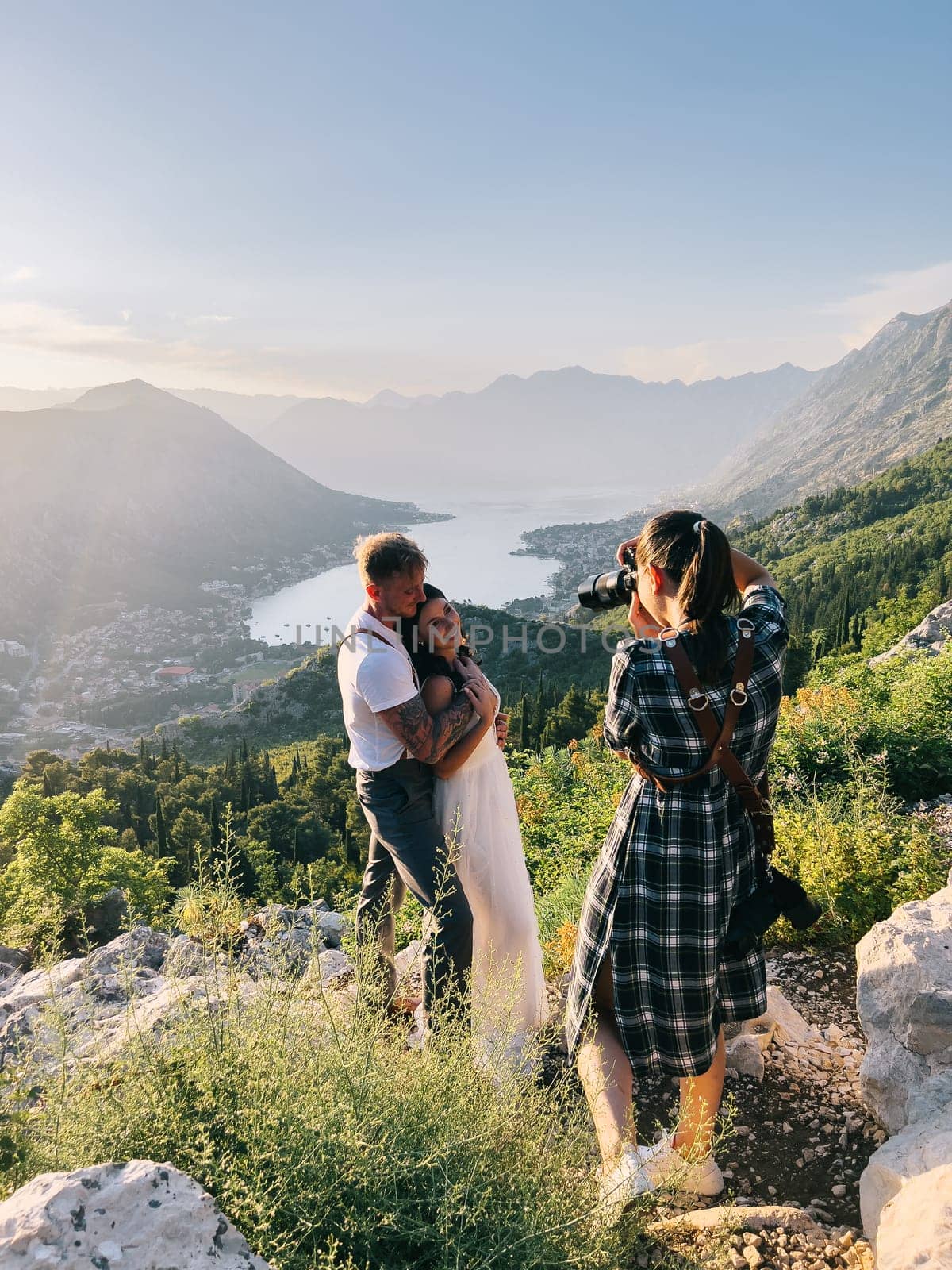 Girl-photographer photographs the embracing bride and groom on a rock above the Kotor Bay. High quality photo