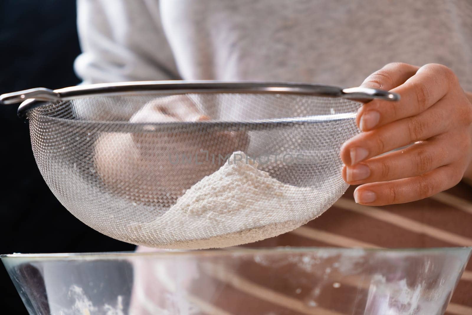 Close up woman sifts wheat flour through a sieve into mixing bowl for dough preparation for cake and bakery on the black background. Knead the dough for homemade pastry.