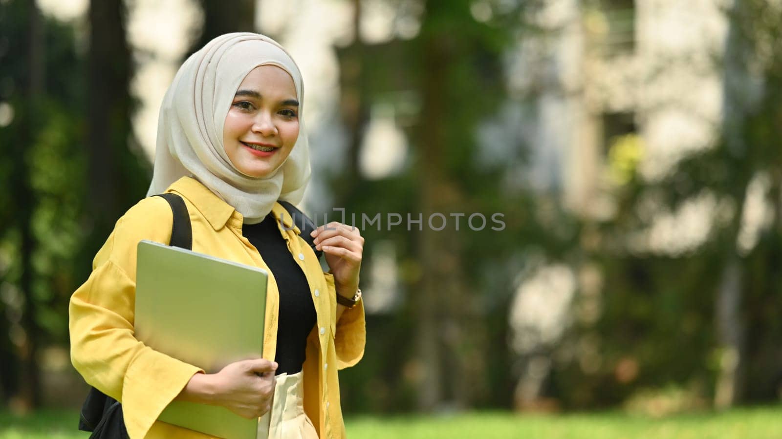 Smiling muslim college student holding laptop standing in the campus. Education, technology and lifestyle concept by prathanchorruangsak