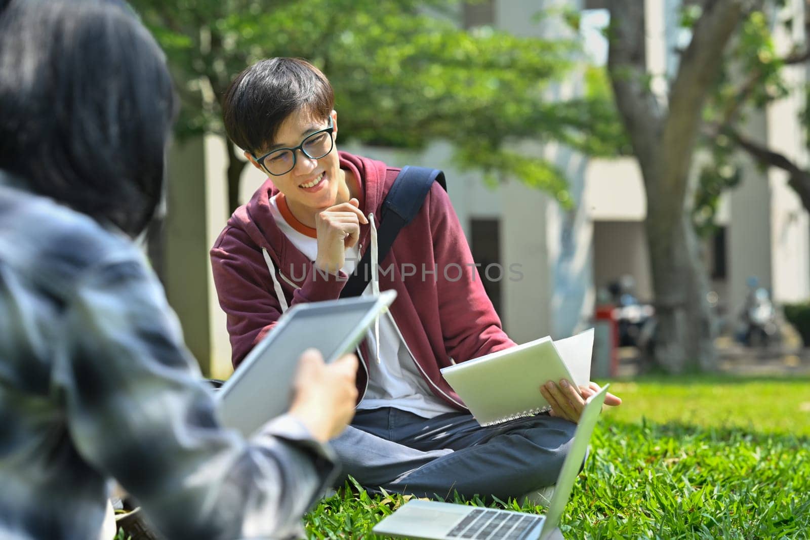 Asian man student reading books, preparing for exam while sitting on green meadow in campus. Education and lifestyle concept.