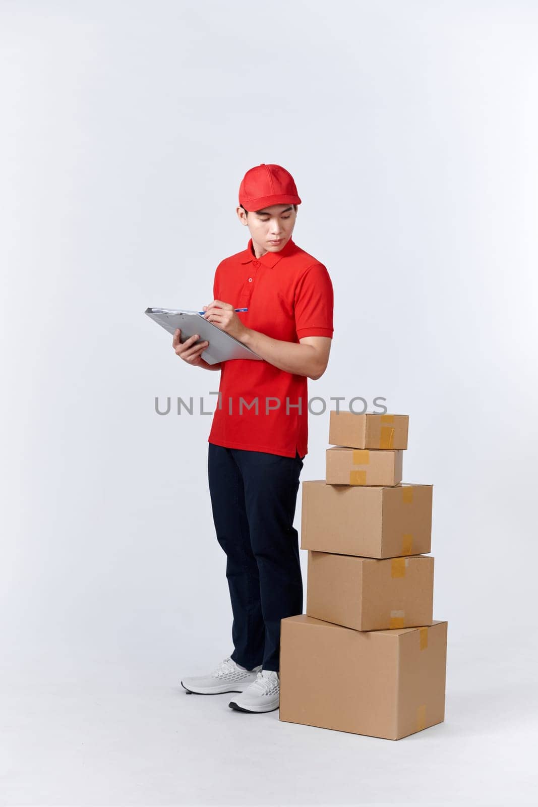 Cheerful professional male mover in casual wear making notes, holding clipboard, standing near a stack of cardboard boxes while doing home relocation. Service concept by makidotvn
