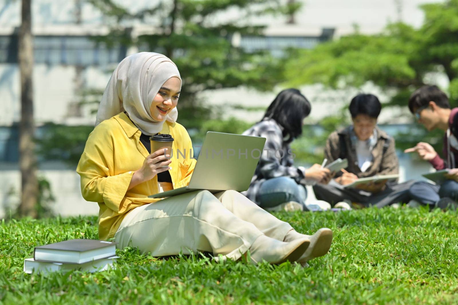 Smiling asian muslim student using laptop on green grass in campus with friends sitting on background by prathanchorruangsak