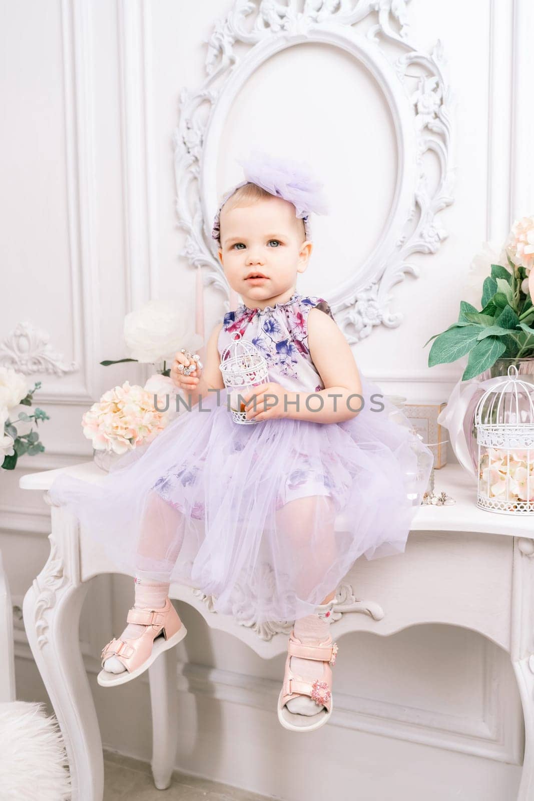 Baby girl elegant dress. A one-year-old girl in a puffy dress and a cute bow poses against the backdrop of a bright room with a dressing table and flowers. by Matiunina