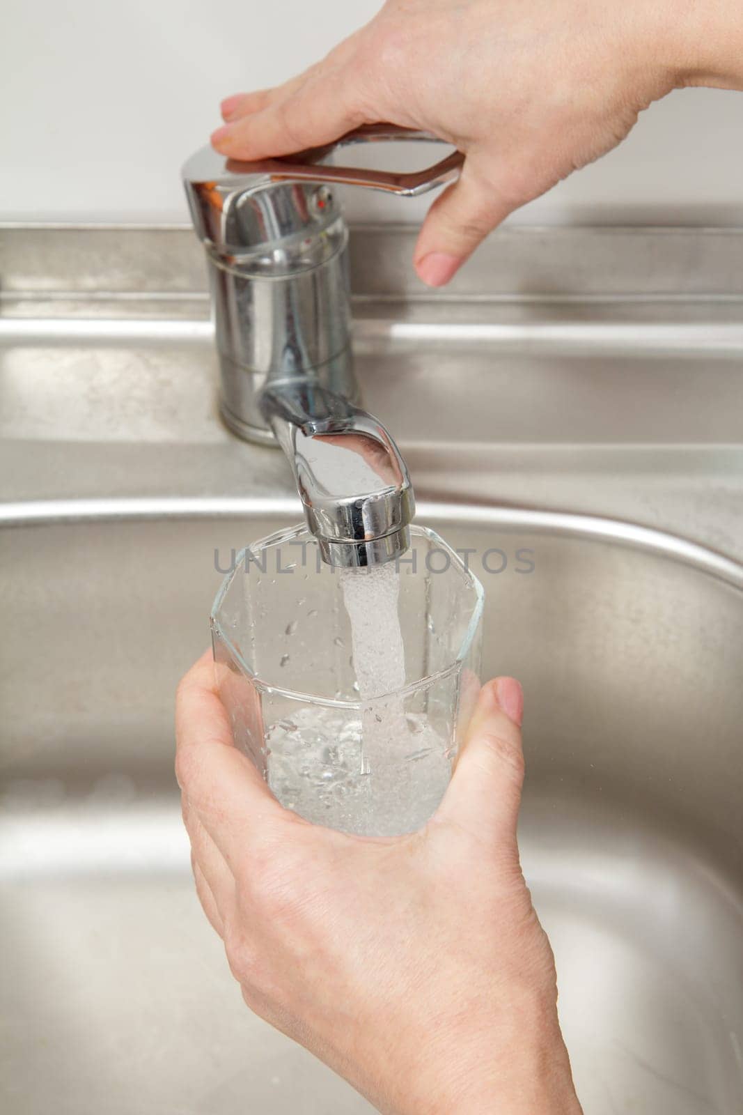 Glass in female hand is filled with clean transparent water under the tap with metal sink on the background