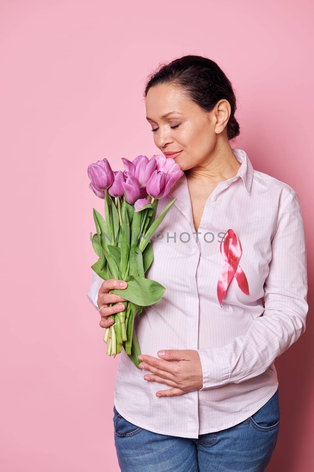 Delightful pregnant woman wearing a pink satin ribbon, sniffing a bouquet of purple tulips, holding her hands on her tummy, isolated pink background. Breast Cancer Awareness Campaign. Female health