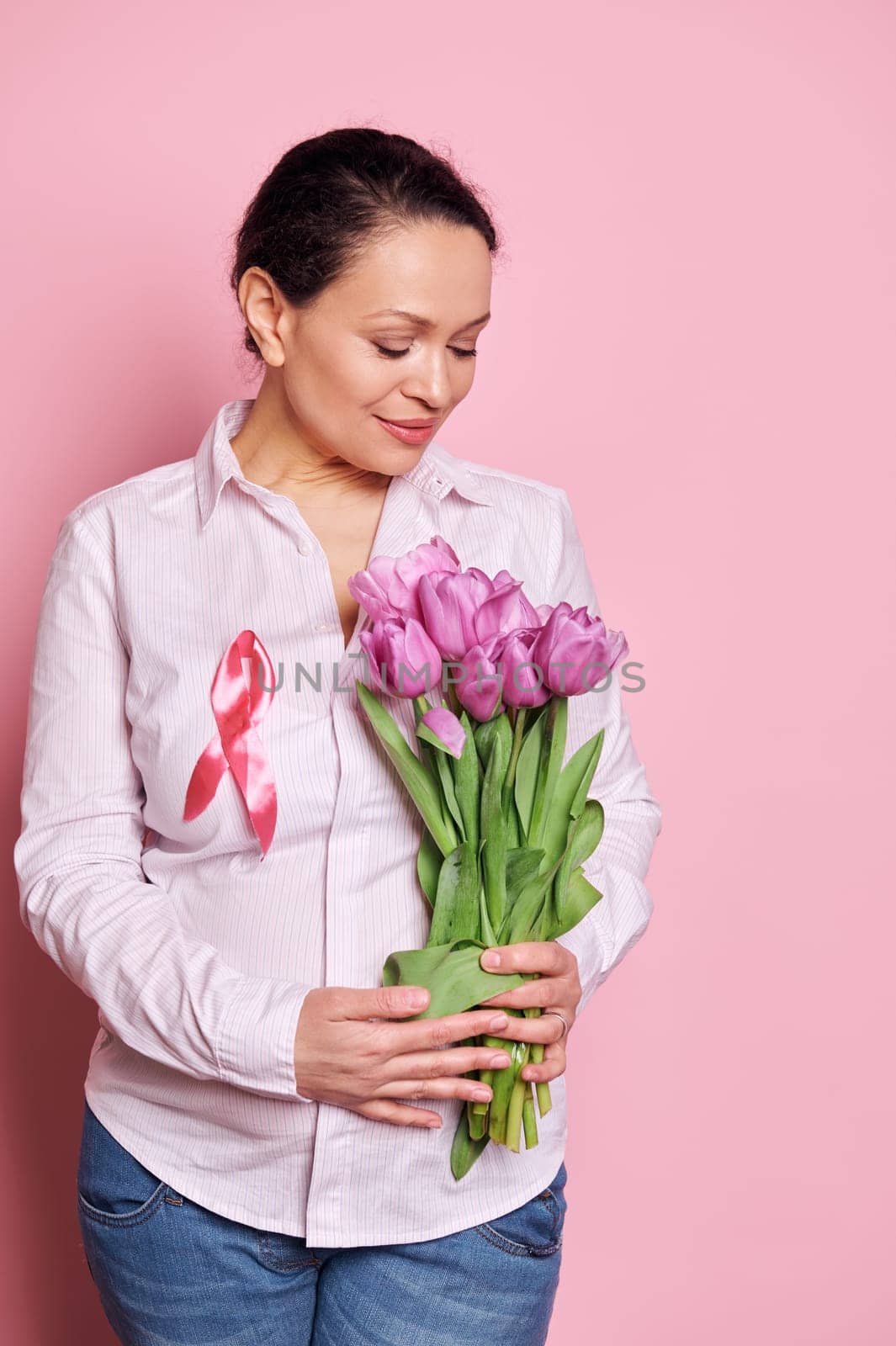Confident portrait on pink background of a pregnant pretty woman, wearing pink satin awareness ribbon, holding purple tulips on isolated color background. Pink October - Breast cancer Awareness month