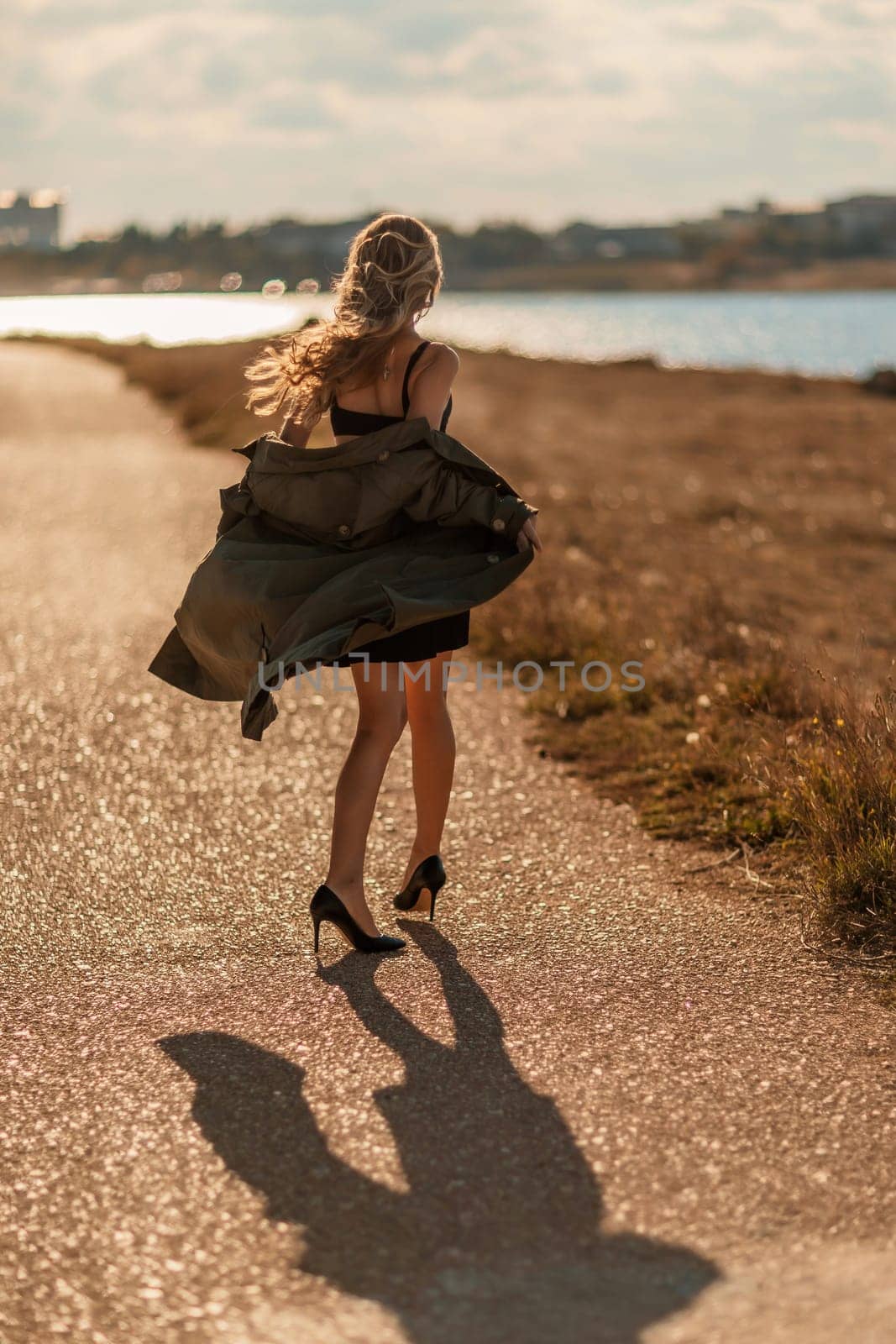 Portrait blonde sea cape. A calm young blonde in an unbuttoned khaki raincoat walks along the seashore, under a raincoat a black skirt and top by Matiunina