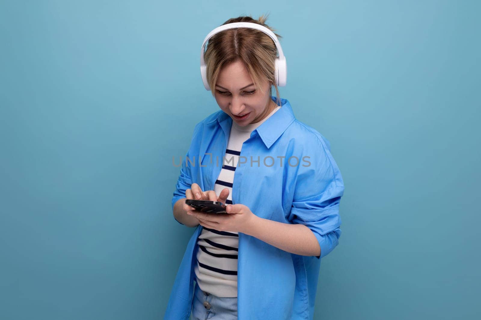 close-up photo of a bright smiling blond young woman dressed in a striped sweater and casual shirt with white wireless headphones communicates in the application in a smartphone on a blue background by TRMK