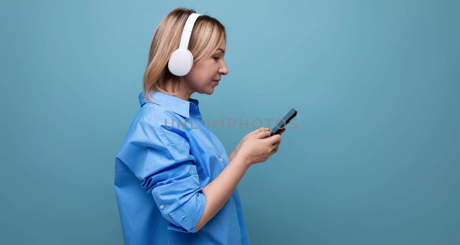 profile horizontal photo of an attentive millennial woman in a casual shirt with large white headphones with a smartphone in her hands on a blue isolated background with copy space by TRMK