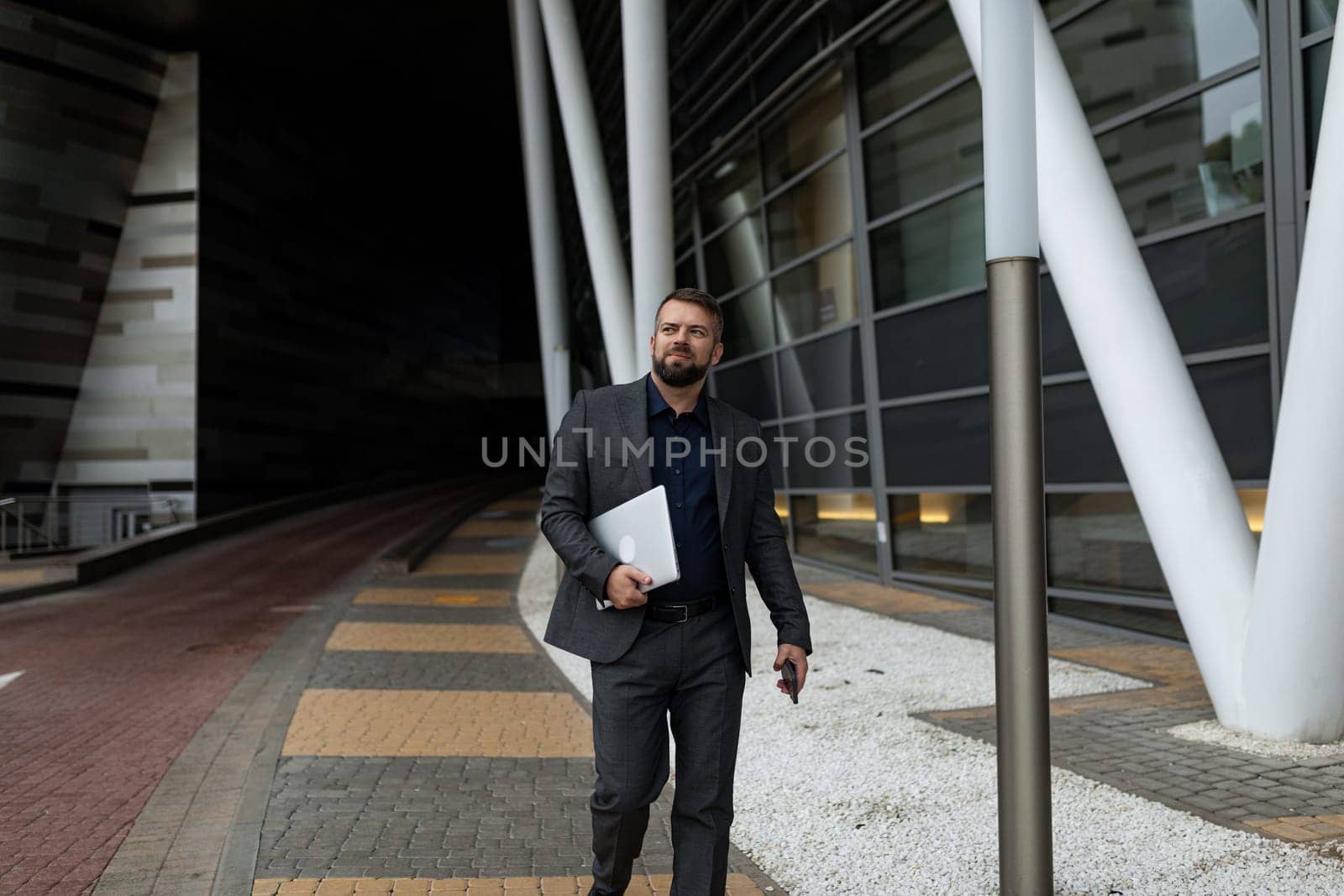 an adult bearded businessman with a laptop in his hands walks outside along the office building.