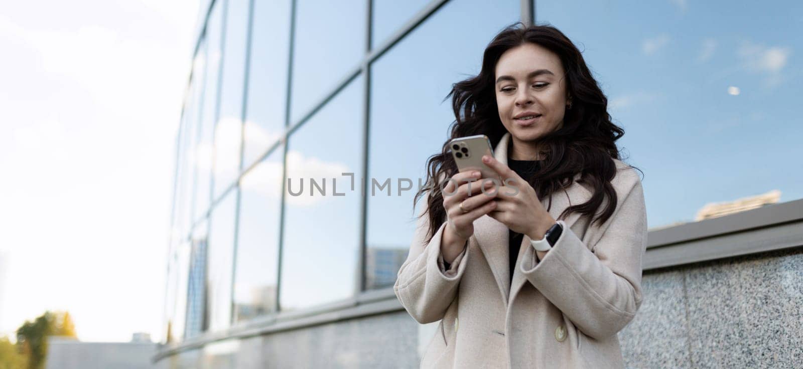 A business woman in her 30s looks at a smartphone screen against the background of a glass facade of a business center.